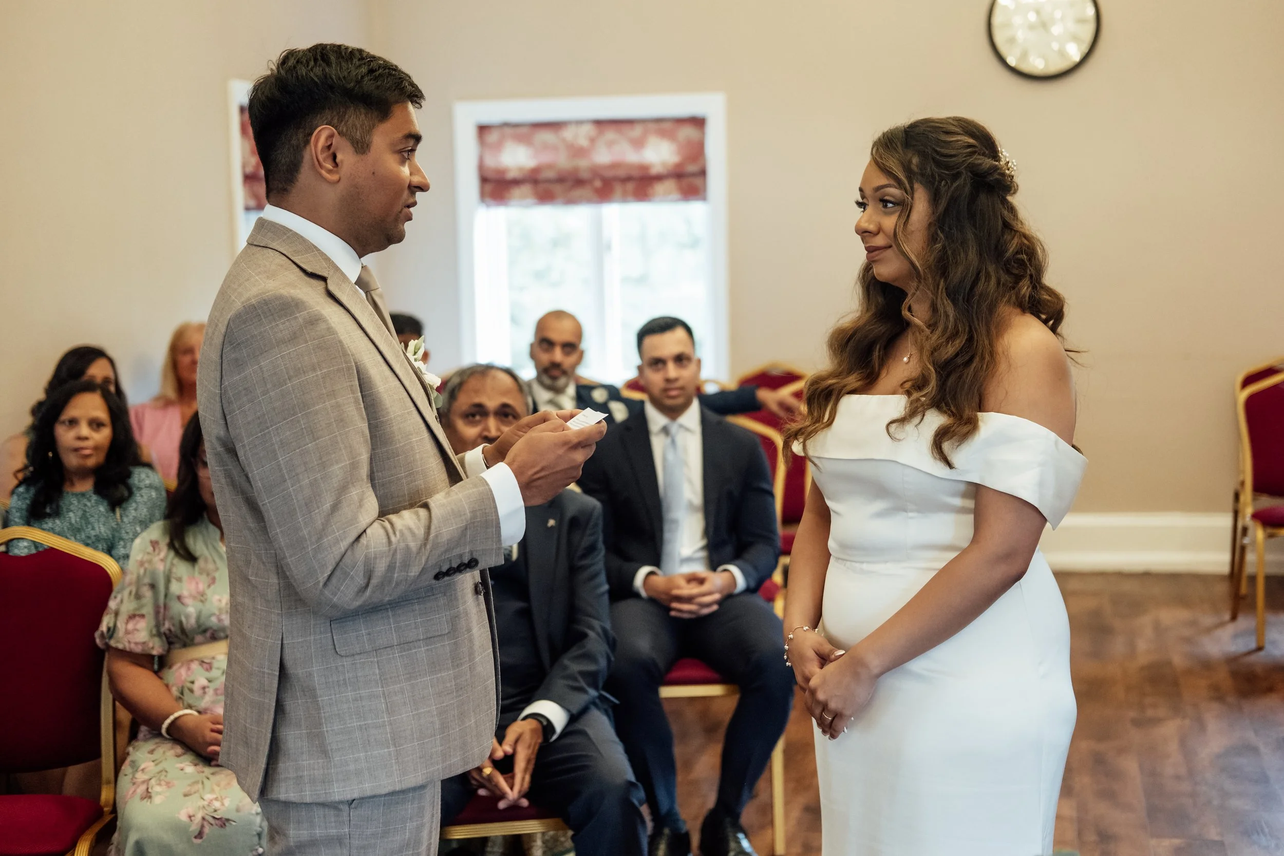 A couple getting married in a small ceremony with guests seated behind them in a room with beige walls and a clock on the wall.