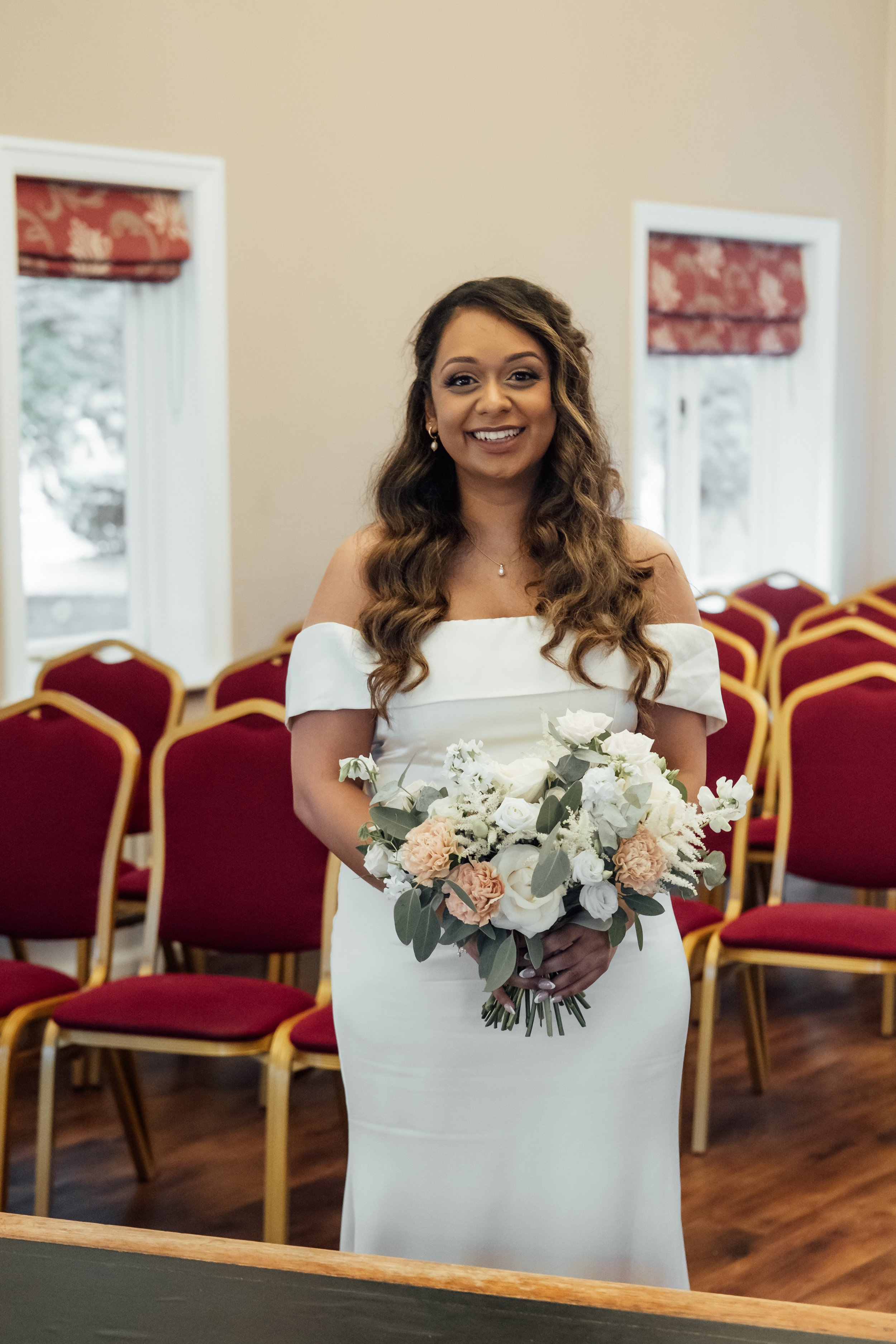 A woman in a white wedding dress holding a bouquet of white and peach flowers, standing inside a room with red chairs and large windows.