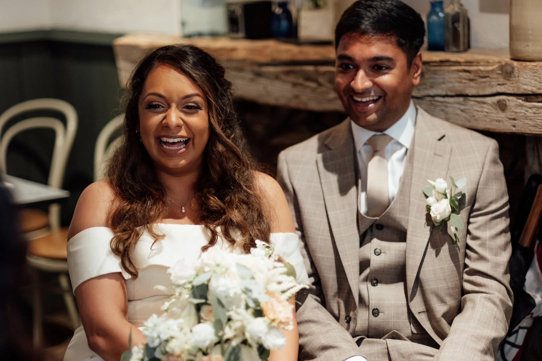 A bride and groom sitting together, smiling and laughing during their wedding ceremony. The bride is holding a bouquet of white flowers, and the groom is wearing a beige suit with a white shirt and a boutonniere.