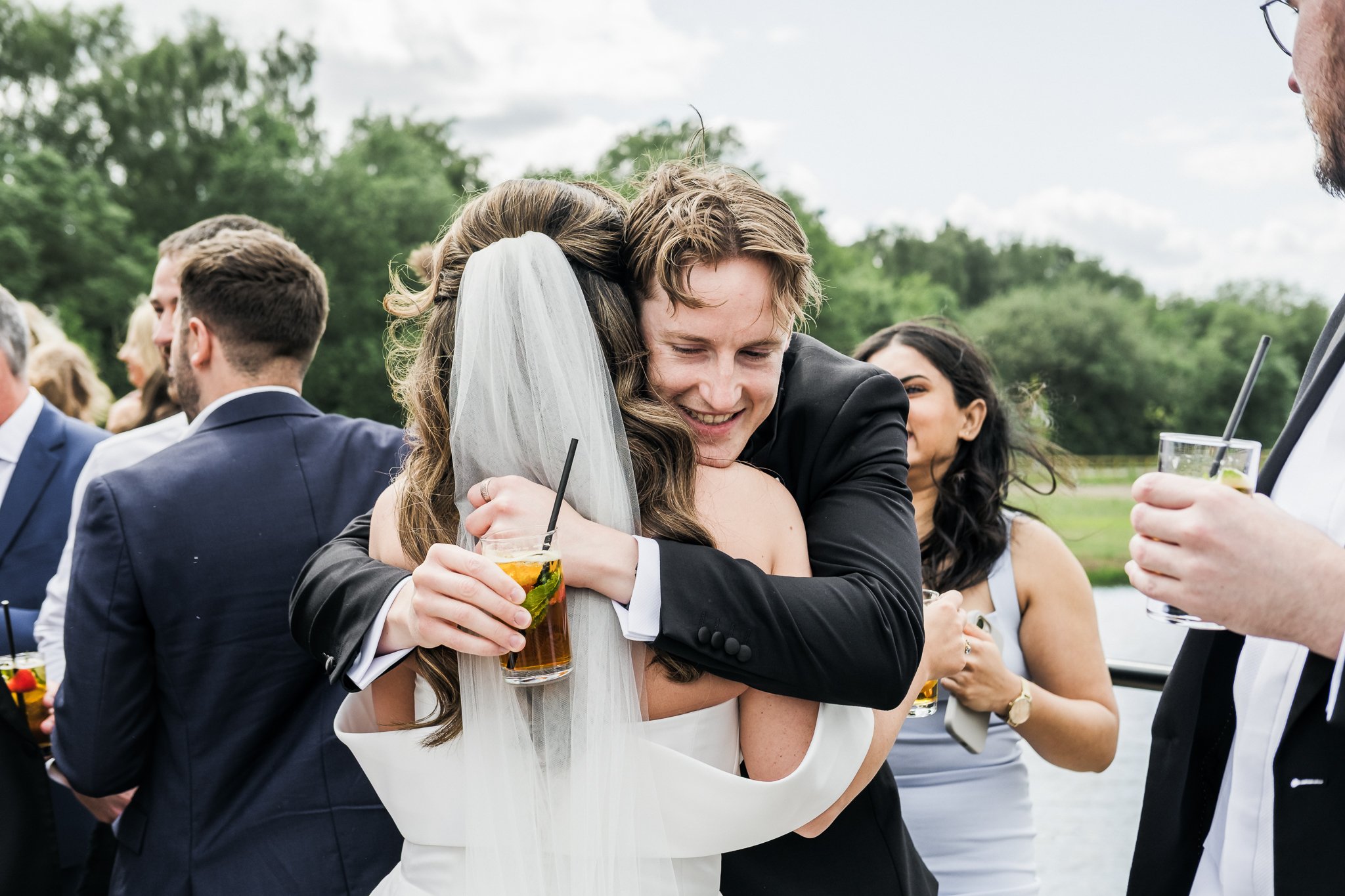 A wedding celebration outdoors with a bride and groom hugging, holding drinks, surrounded by friends.