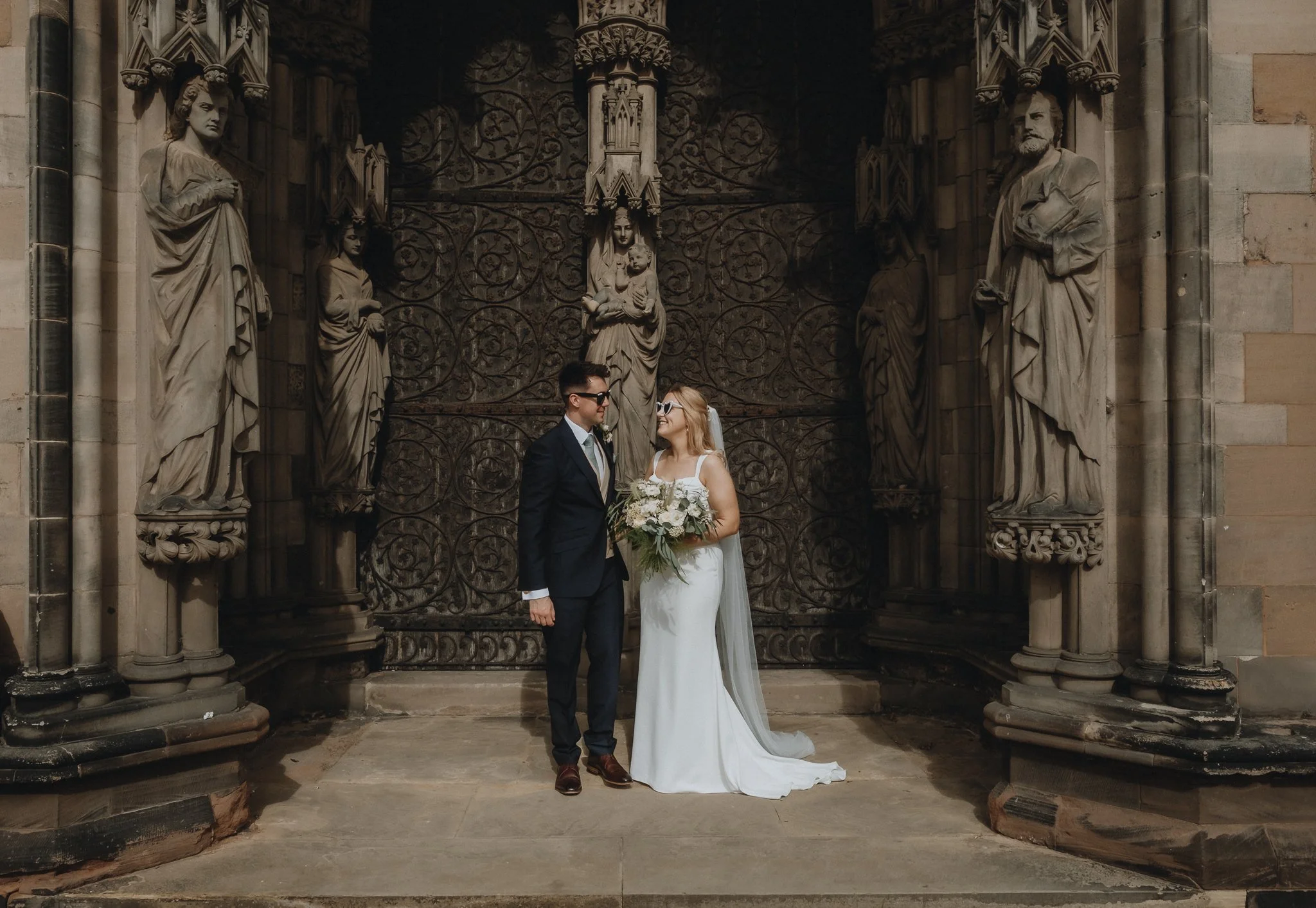 A bride and groom standing in front of an ornate gothic church door with statues and intricate metalwork, on their wedding day.