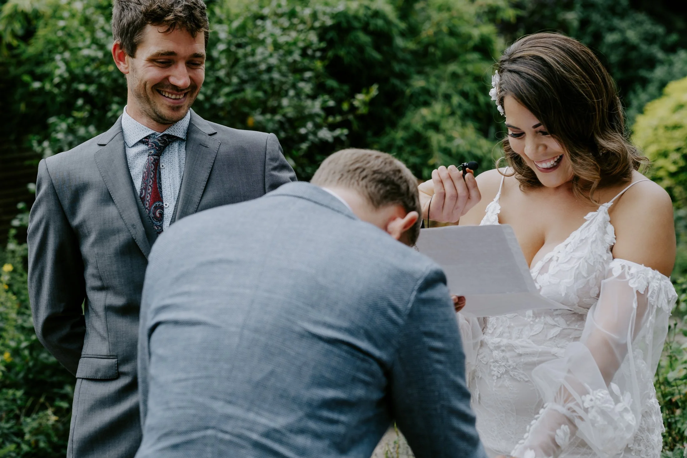 A bride and groom are exchanging vows outdoors, with a woman holding a microphone and reading from a paper, and a man standing beside them, all smiling amid lush greenery.