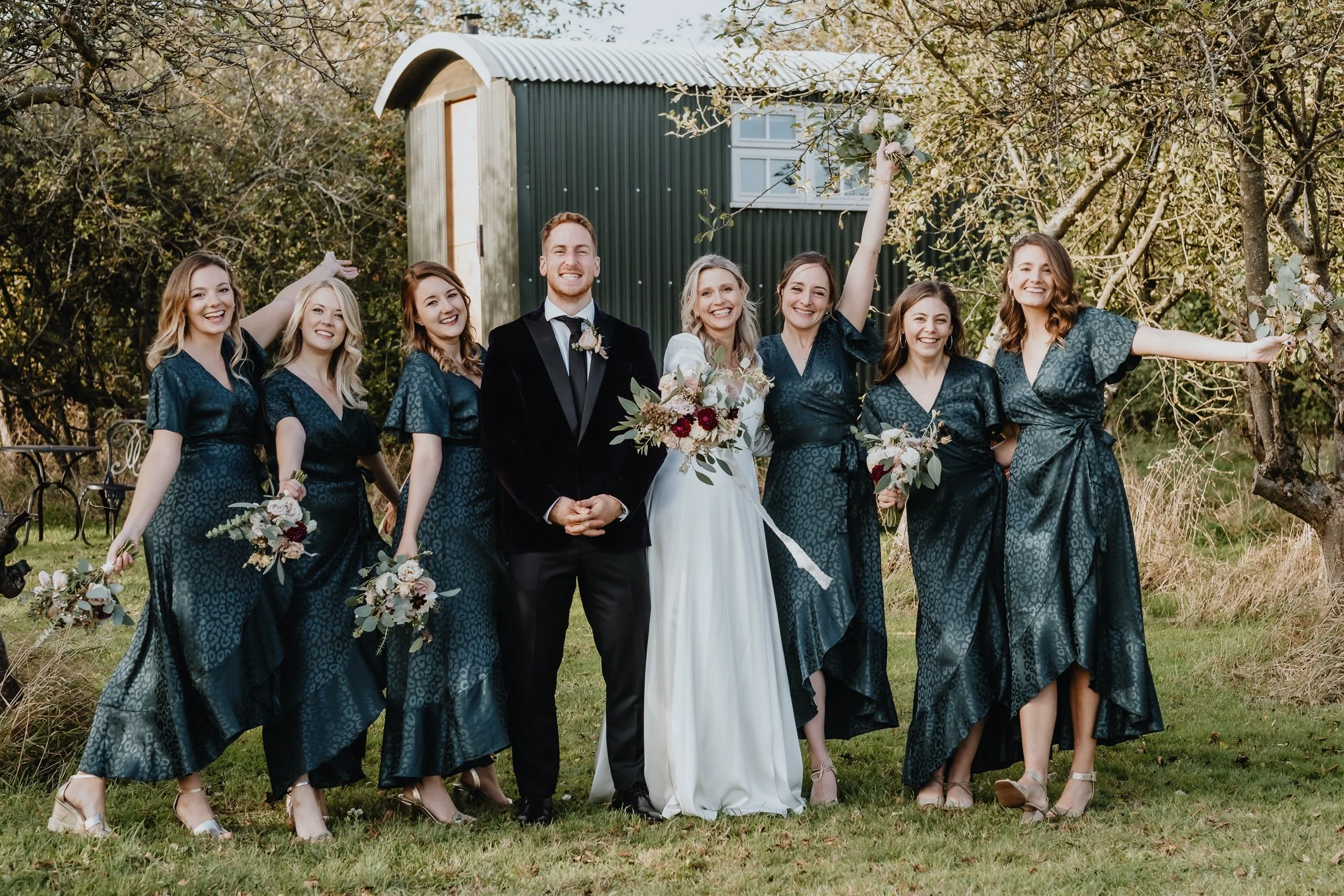 A group of nine people at a wedding outdoors, including a bride in a white dress holding a bouquet, a groom in a black tuxedo, and seven bridesmaids in matching dark teal dresses holding bouquets, standing in front of a green shed and trees.