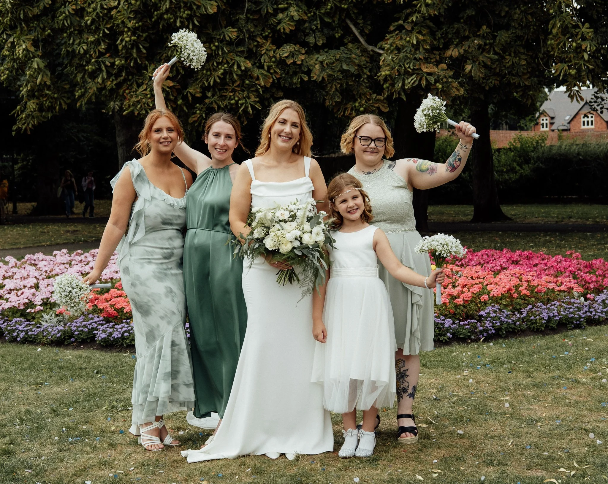 Group of five women and one girl smiling at a wedding, holding flowers, outdoors in front of a flower bed and trees.