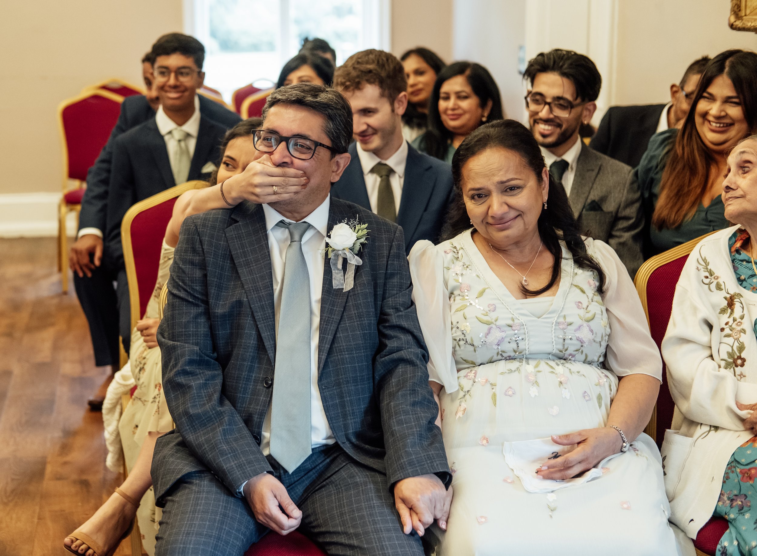 Group of people attending a wedding ceremony, sitting on red chairs. The groom, wearing glasses and a checked suit with a white flower on his lapel, is covering his mouth and appears emotional. The bride, sitting next to him in a white dress with flo