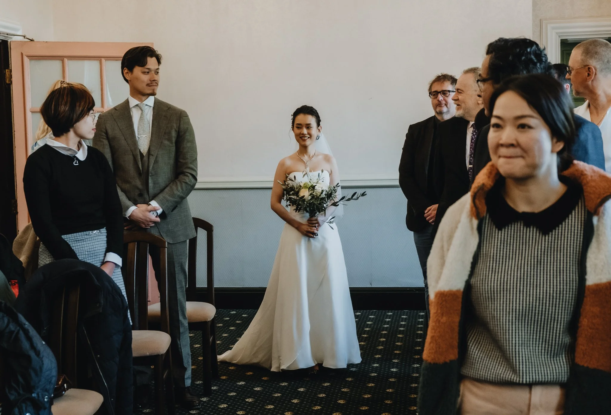 A wedding scene with a bride in a strapless white gown holding a bouquet of flowers, standing in the center surrounded by guests, in a room with light-colored walls and dark patterned carpet.