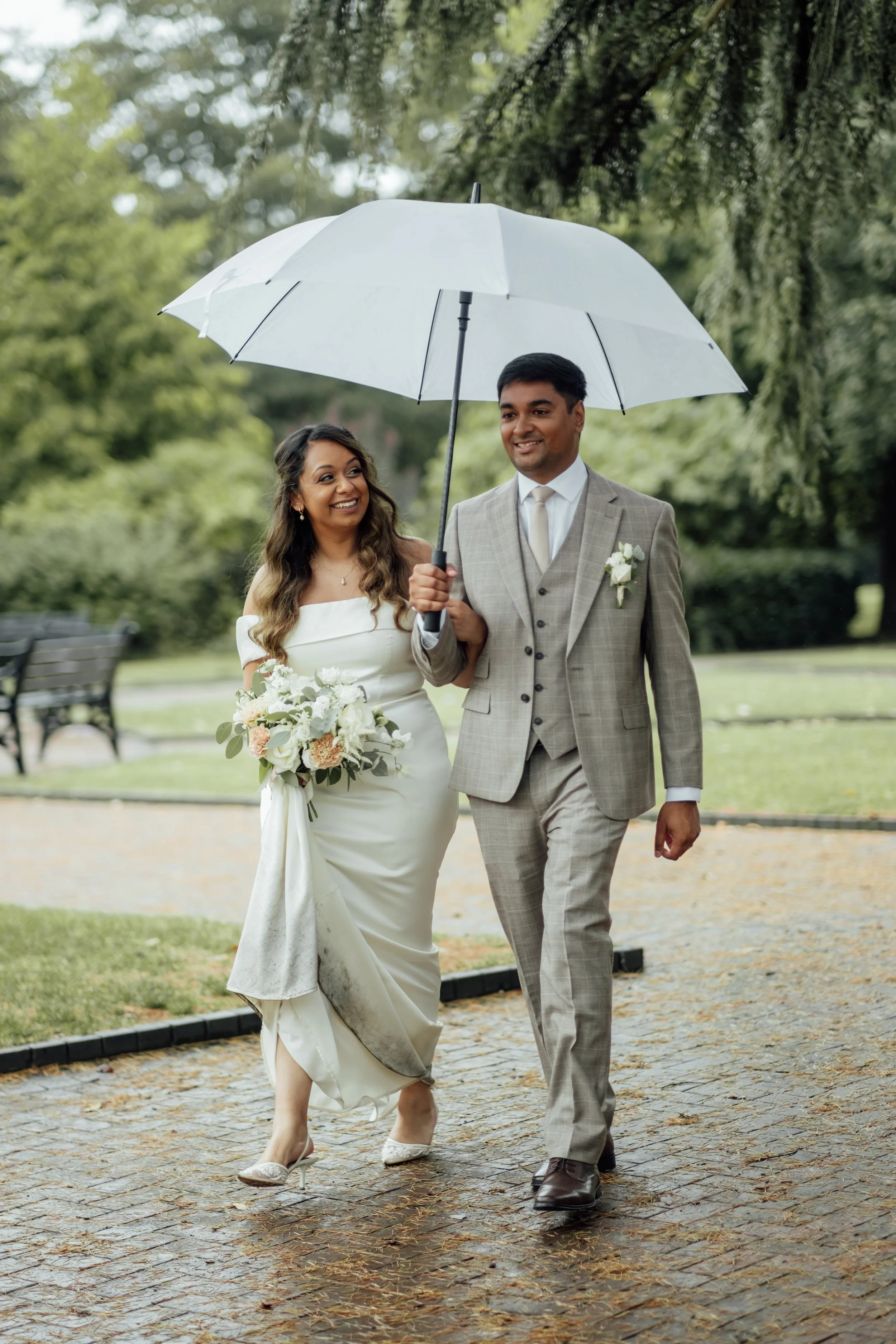 A newlywed couple walking outdoors on a rainy day, sharing a white umbrella, with the bride holding a bouquet of white flowers and wearing a white wedding gown, and the groom in a light gray suit at Bridgford Hall.