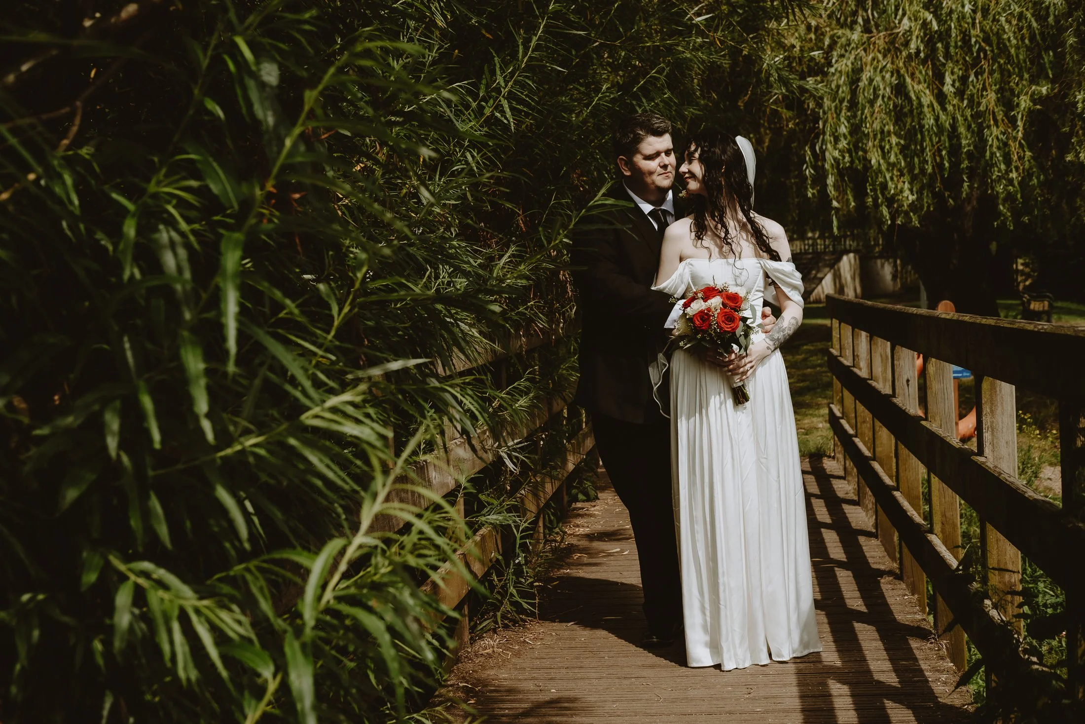 A bride and groom standing closely on a wooden pathway surrounded by greenery, with the bride holding a bouquet of red roses, during their wedding photoshoot.