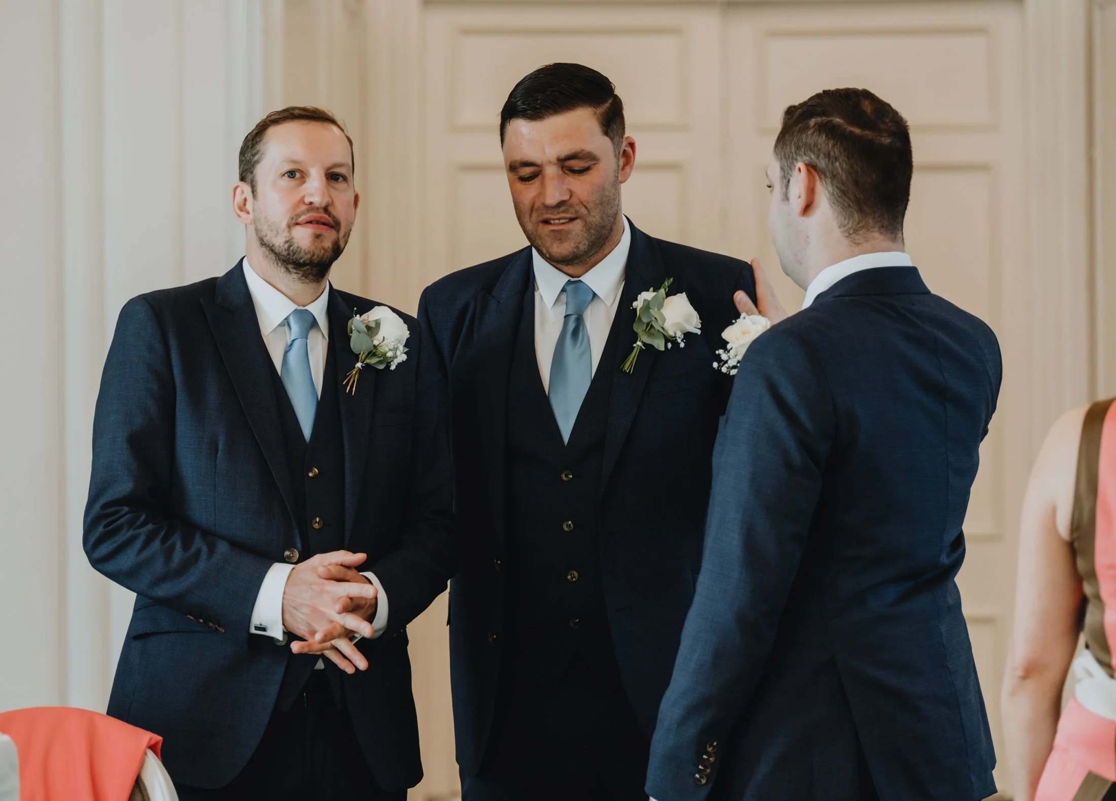 Three men in suits with boutonnières attending a formal event, standing close together and engaging in conversation inside a room with wood-paneled walls.