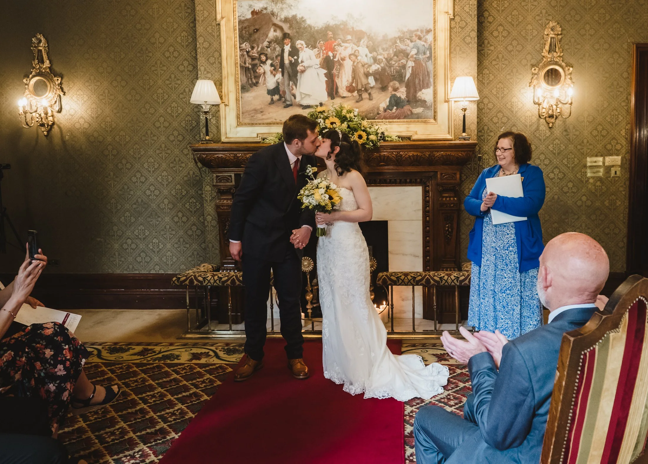 A bride and groom kiss during their wedding ceremony in a richly decorated room with antique wallpaper, a large framed painting, and a marble fireplace. The bride is holding a bouquet of flowers, and a woman in blue stands nearby holding a folder, sm