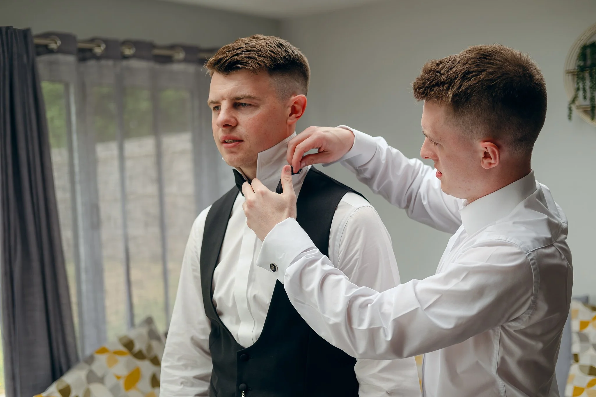 Two men in formal attire, one helping the other adjust his collar or bow tie in a room with large windows and curtains