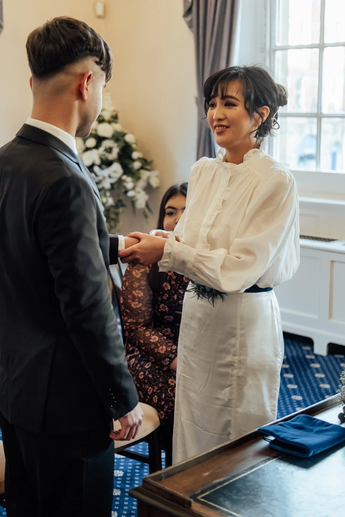 A woman in a white dress holding hands with a man in a black suit during a formal or wedding ceremony, with another woman seated behind them and a window in the background.