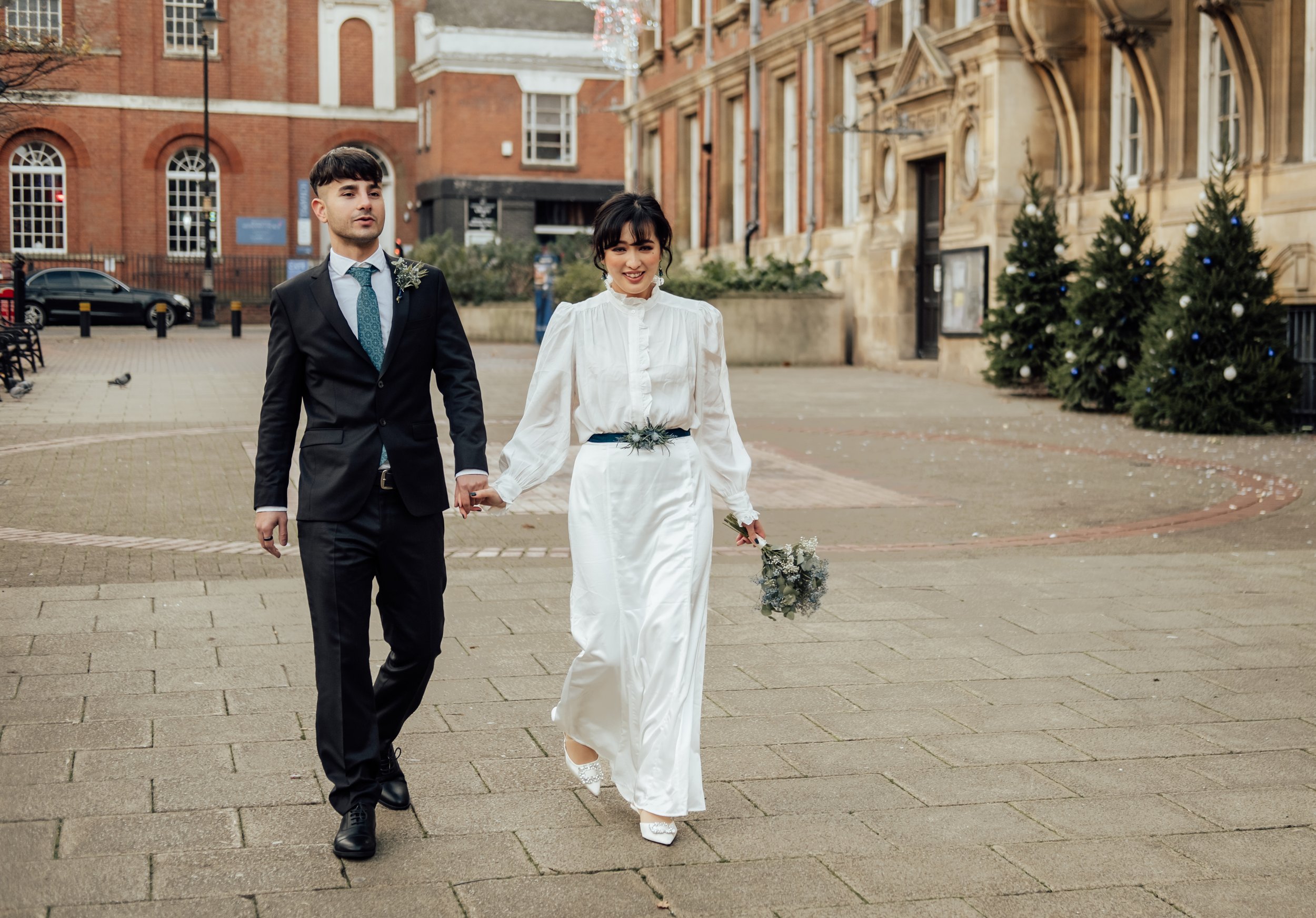A newlywed couple walking hand in hand outside in a city square, with Christmas trees in the background.