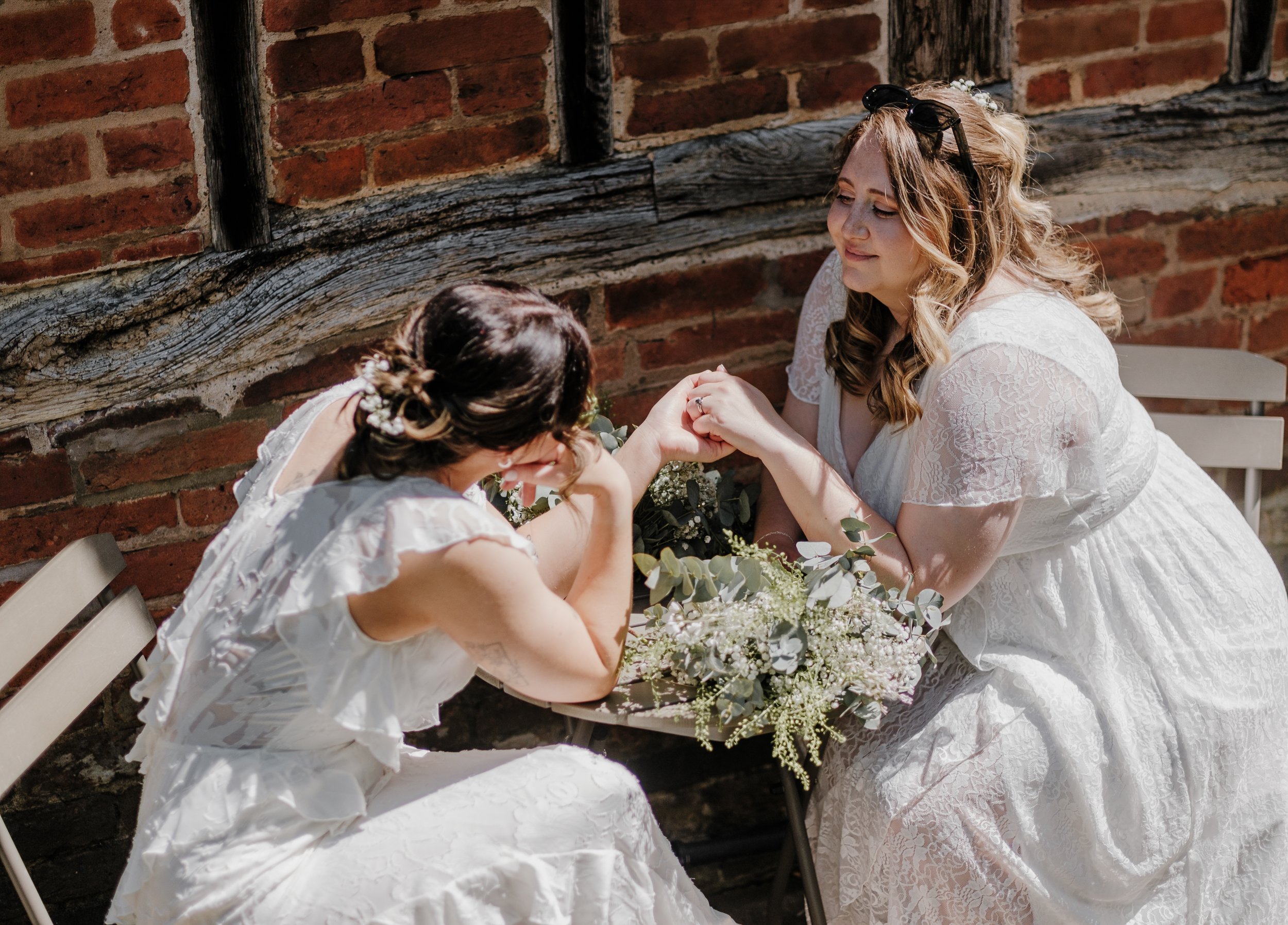 A woman in a white lace dress holding hands with her wife in a white dress, both sitting at a table with a floral centerpiece, against a brick wall.
