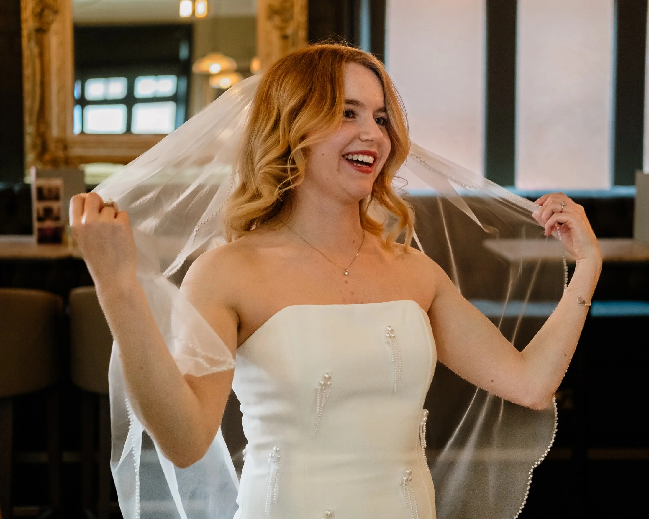 A woman with red hair in a white strapless wedding dress holding a veil, smiling in a warmly lit room with wooden and dark decor elements.