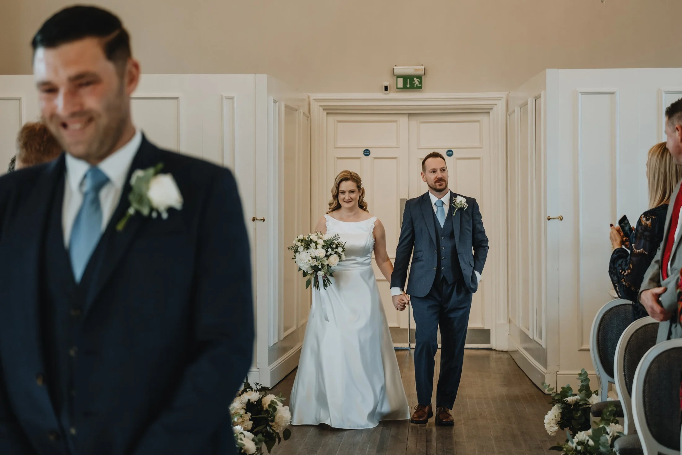 A bride walking down the aisle, with guests on either side, in a wedding ceremony. The bride is in a white dress and holding a bouquet, and the groom is in a dark suit with a boutonniere.