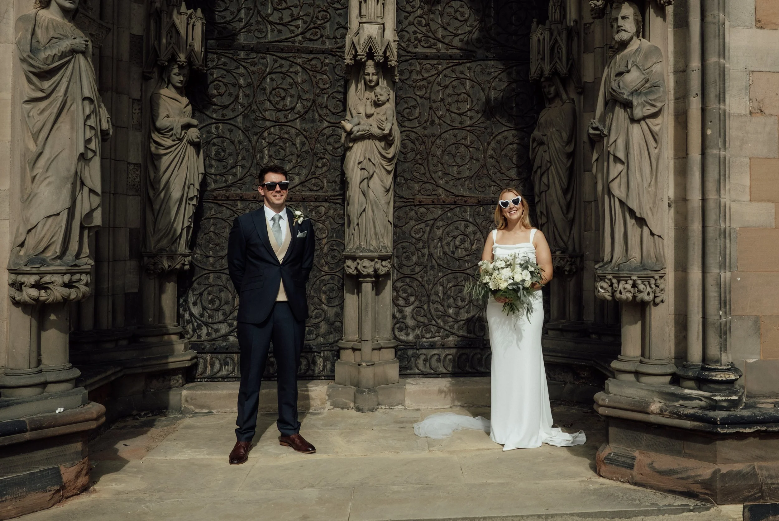 A bride and groom stand in front of an ornate stone church door with sculpted statues and intricate metalwork. The bride wears a white wedding dress and holds a bouquet, while the groom wears a navy suit with a boutonniere and sunglasses.
