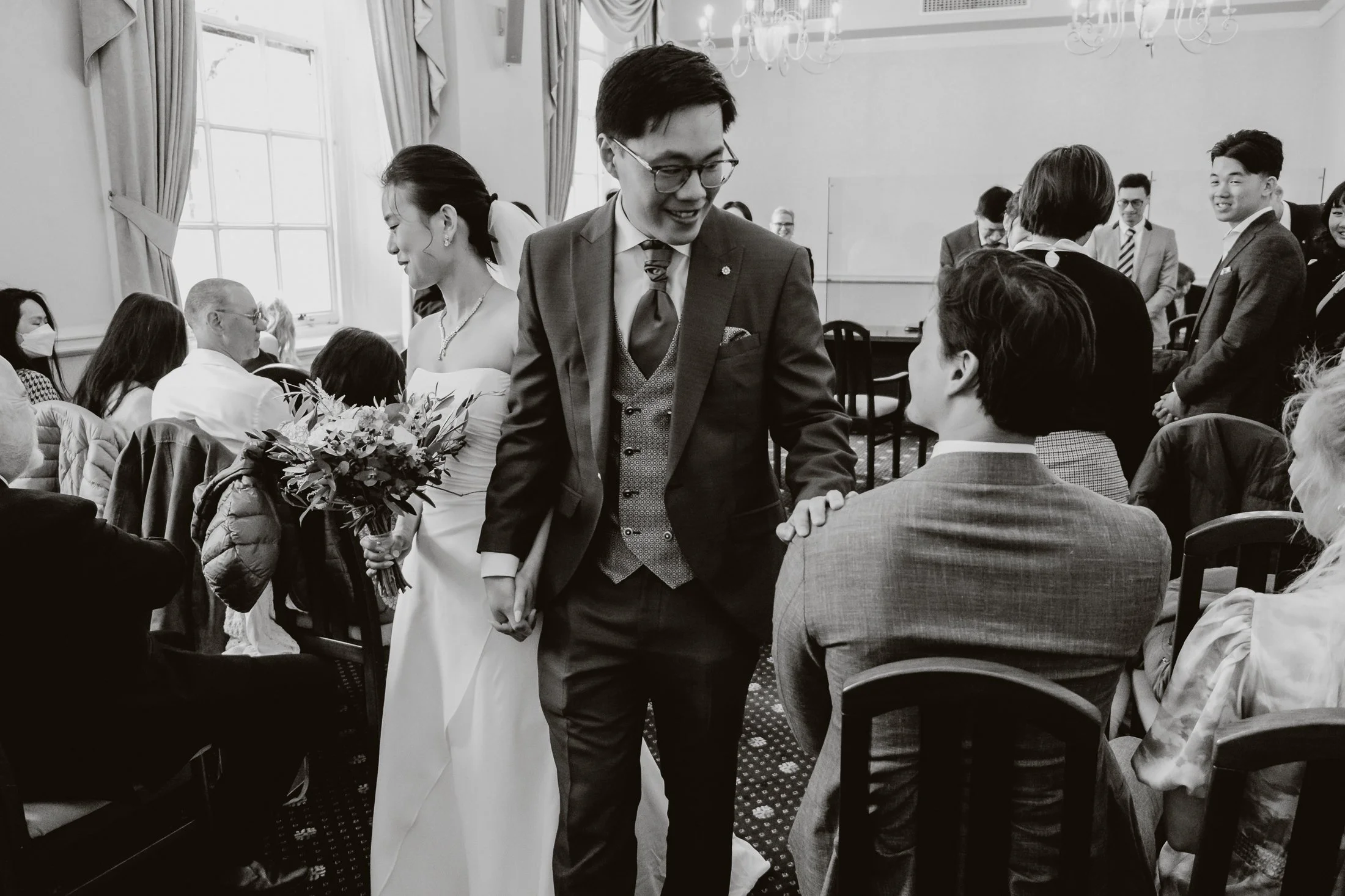 Black and white photo of a wedding ceremony in a decorated room with guests, including a bride with a bouquet and men in suits, engaging in conversation and greeting each other.