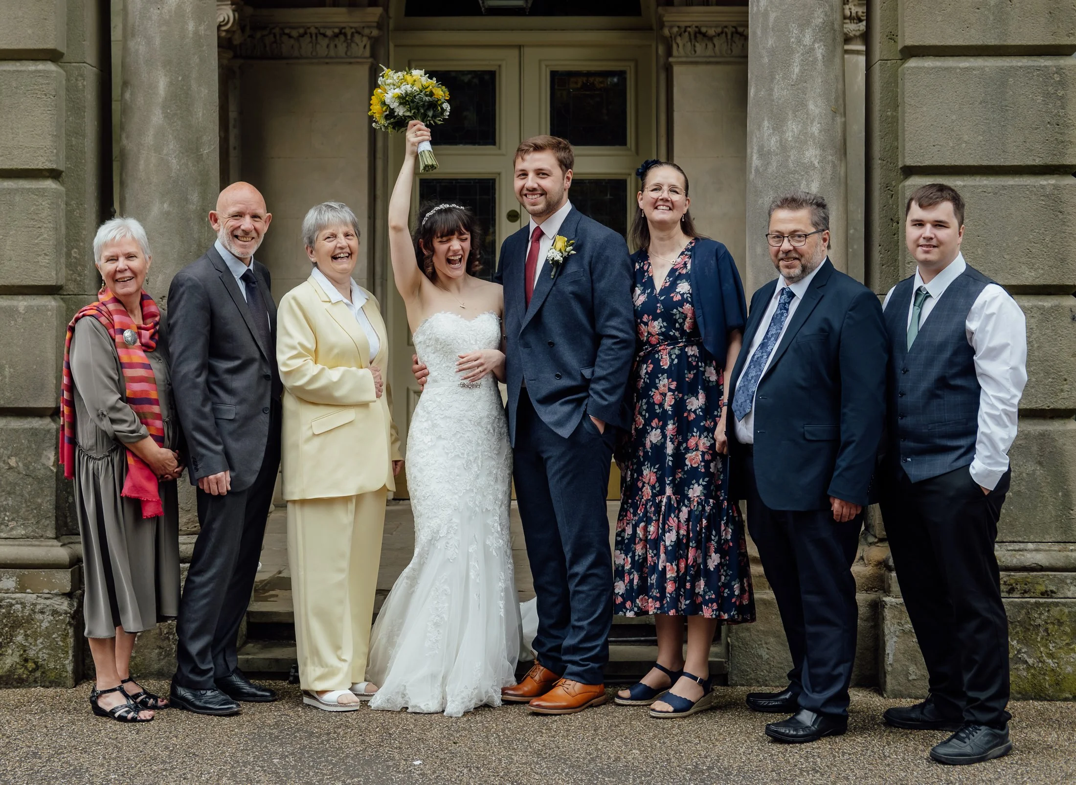 Group of people, including bride and groom, standing outside a building with columns on a wedding day. The bride is holding a bouquet up in the air and the groom is smiling.