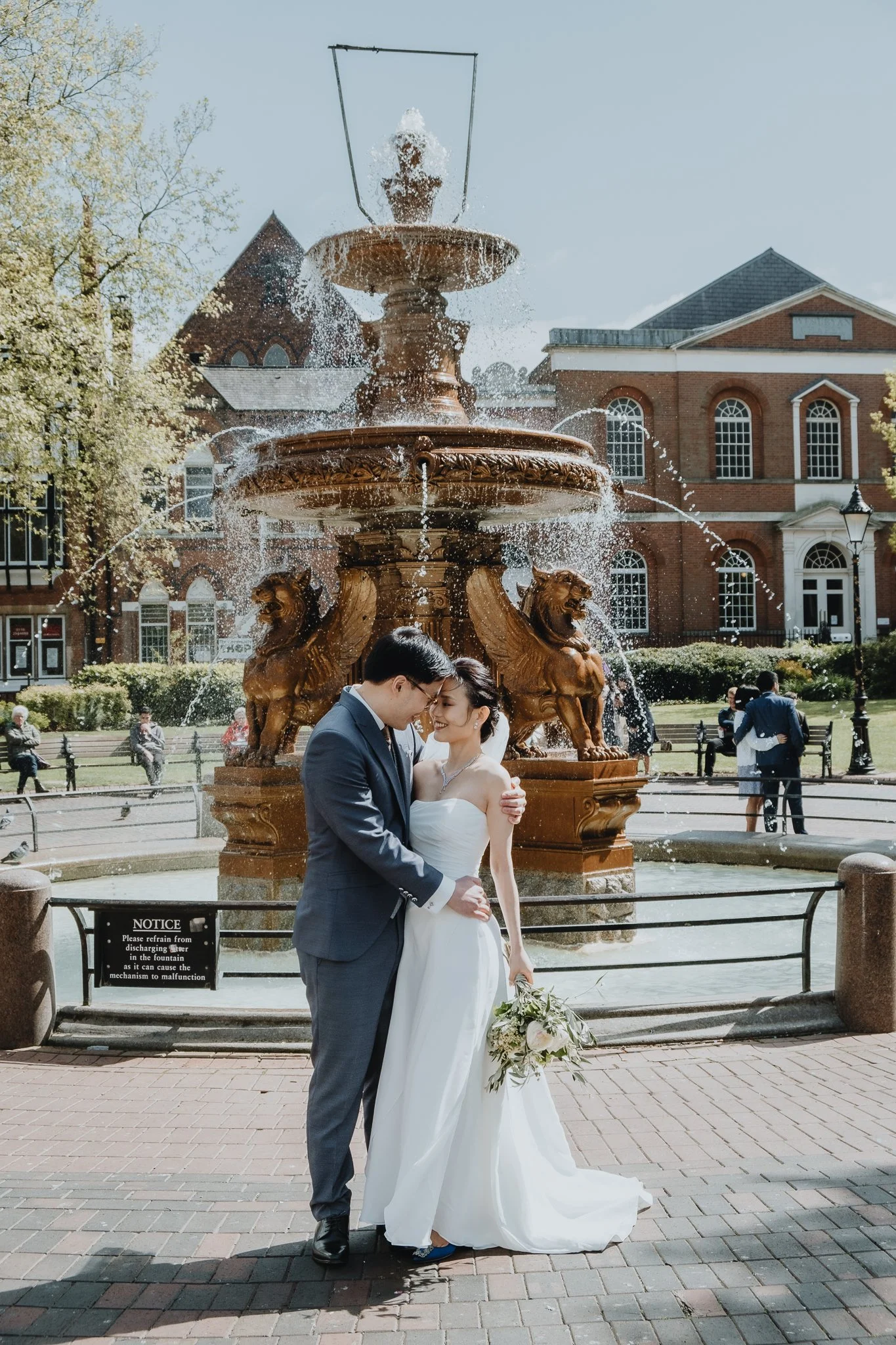 A newlywed couple in wedding attire standing close together in front of a large decorative fountain in an urban park, with people in the background and a historic brick building.