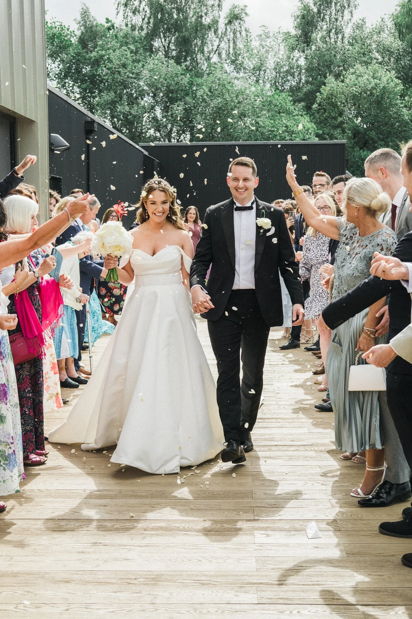 Bride and groom walking hand in hand down an outdoor wooden aisle, surrounded by guests throwing flower petals, on a sunny day with trees in the background.