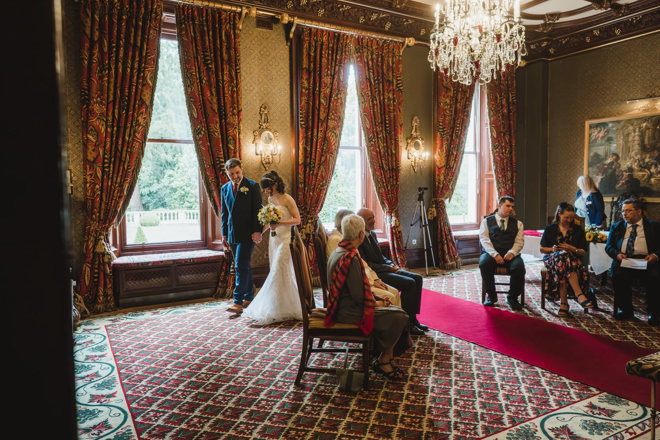 A wedding ceremony in an elegant, richly decorated room with large windows, red and gold drapes, a chandelier, and ornate wall sconces. The bride, dressed in a white gown, and the groom, in a dark suit, are holding hands. Several seated guests, inclu