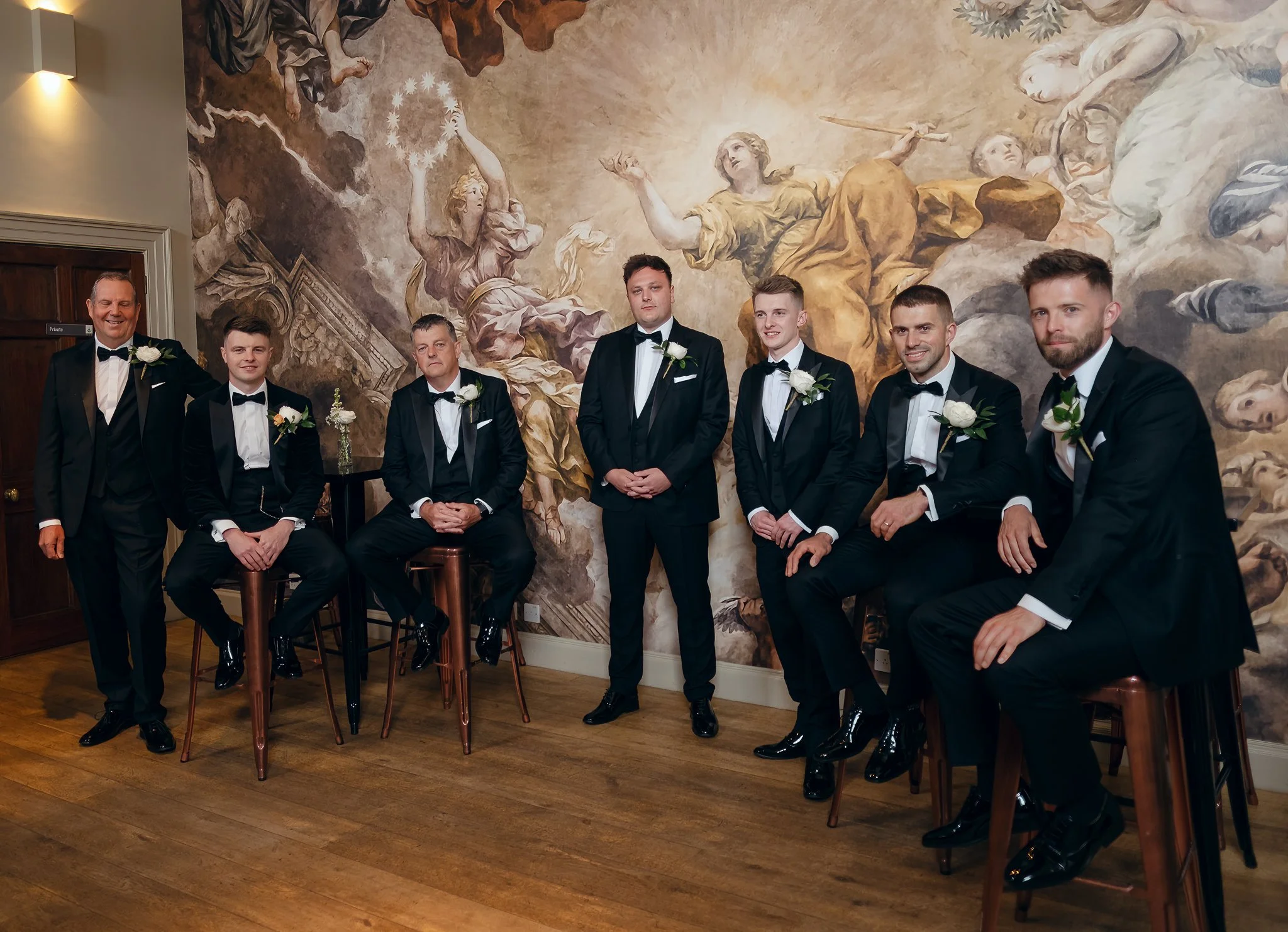Group of seven men dressed in black tuxedos with white shirts and black bow ties, some with white boutonnieres, posed in front of a large baroque-style mural in an indoor setting.