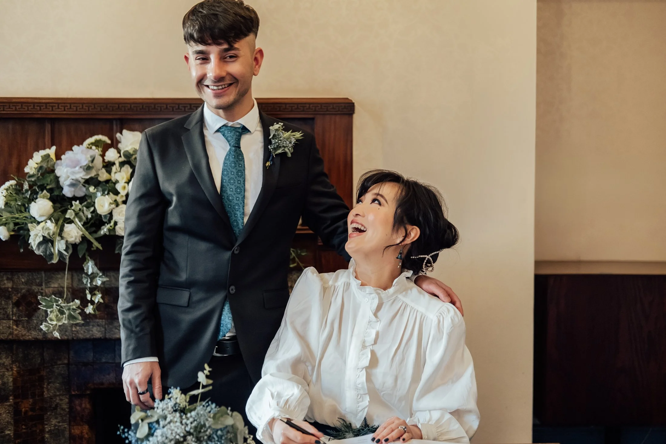A joyful man in a black suit with a teal tie stands next to a seated woman wearing a white blouse, both smiling during a wedding celebration. The man has a boutonniere on his suit lapel, and there are flowers on a table in front of them. The backgrou