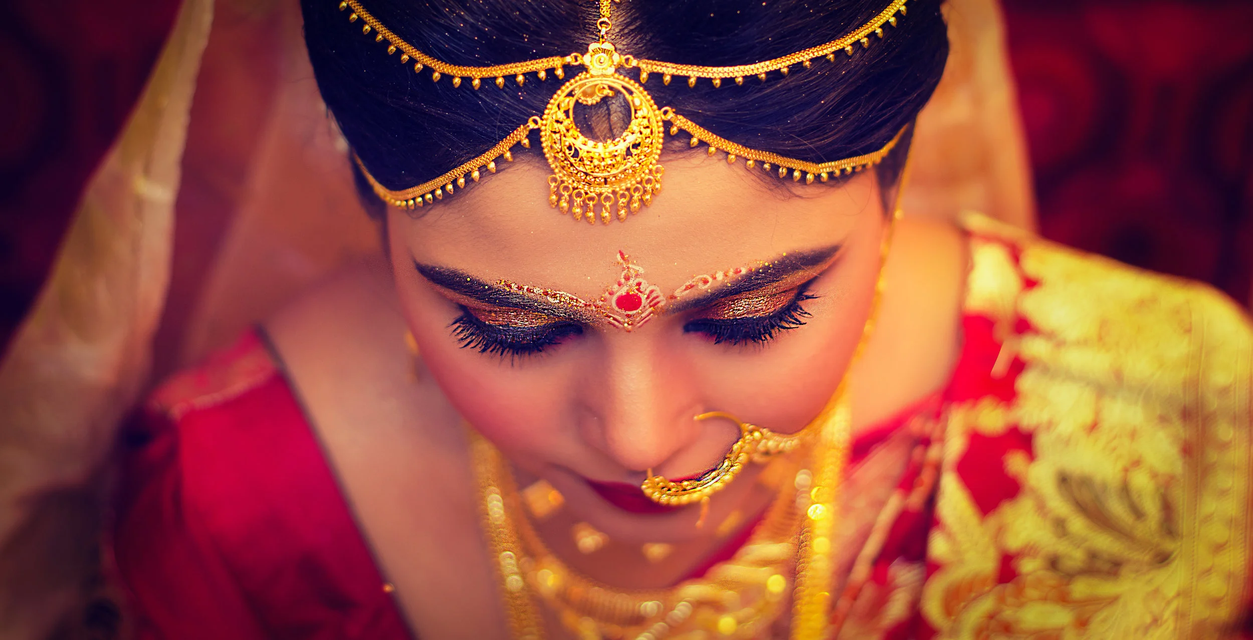 Close-up of a woman dressed in traditional Indian attire with intricate gold jewelry and makeup, bowing her head down.