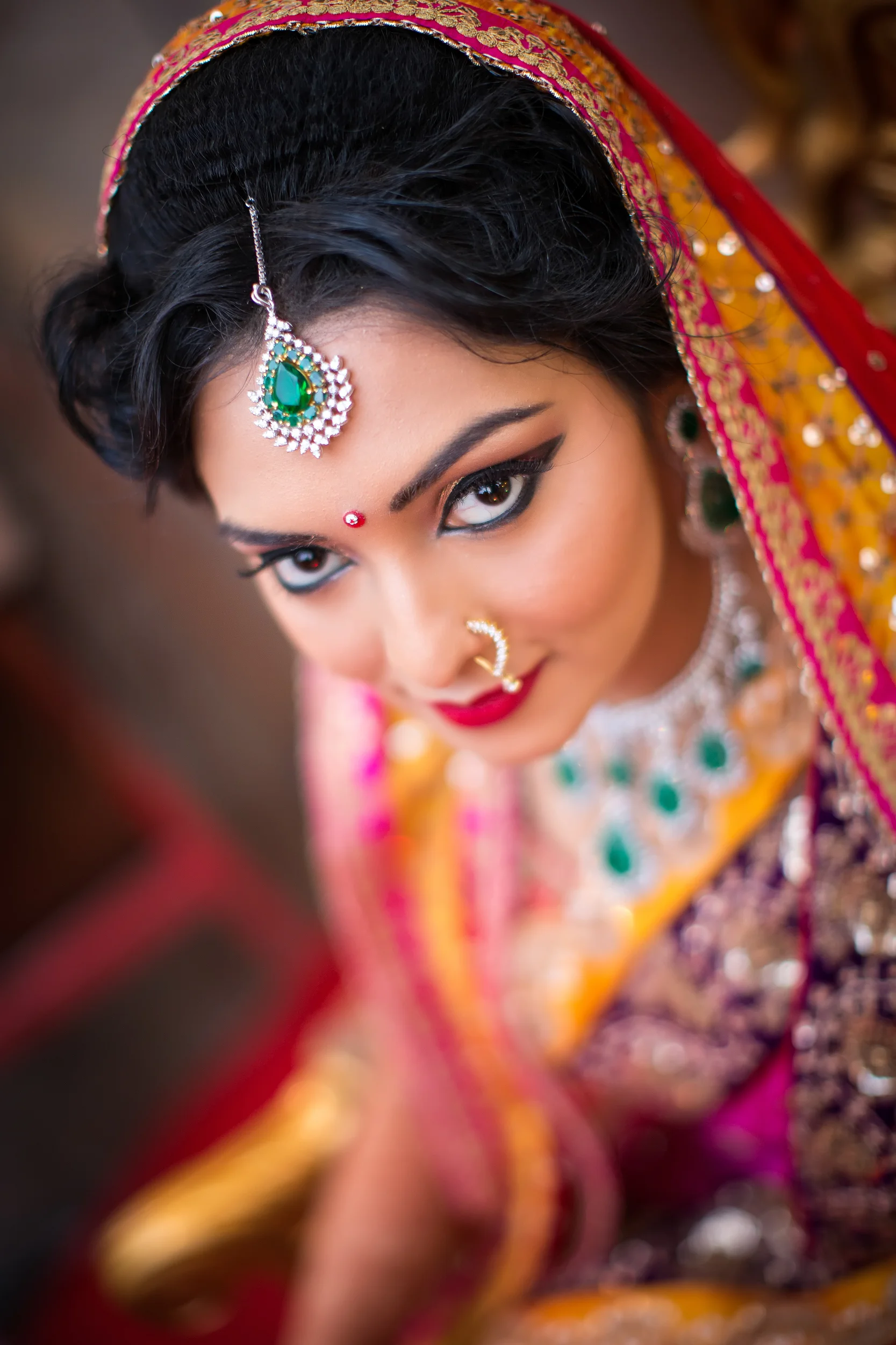 Close-up portrait of a woman wearing traditional Indian attire, jewelry, and makeup, looking up towards the camera with a slight smile.