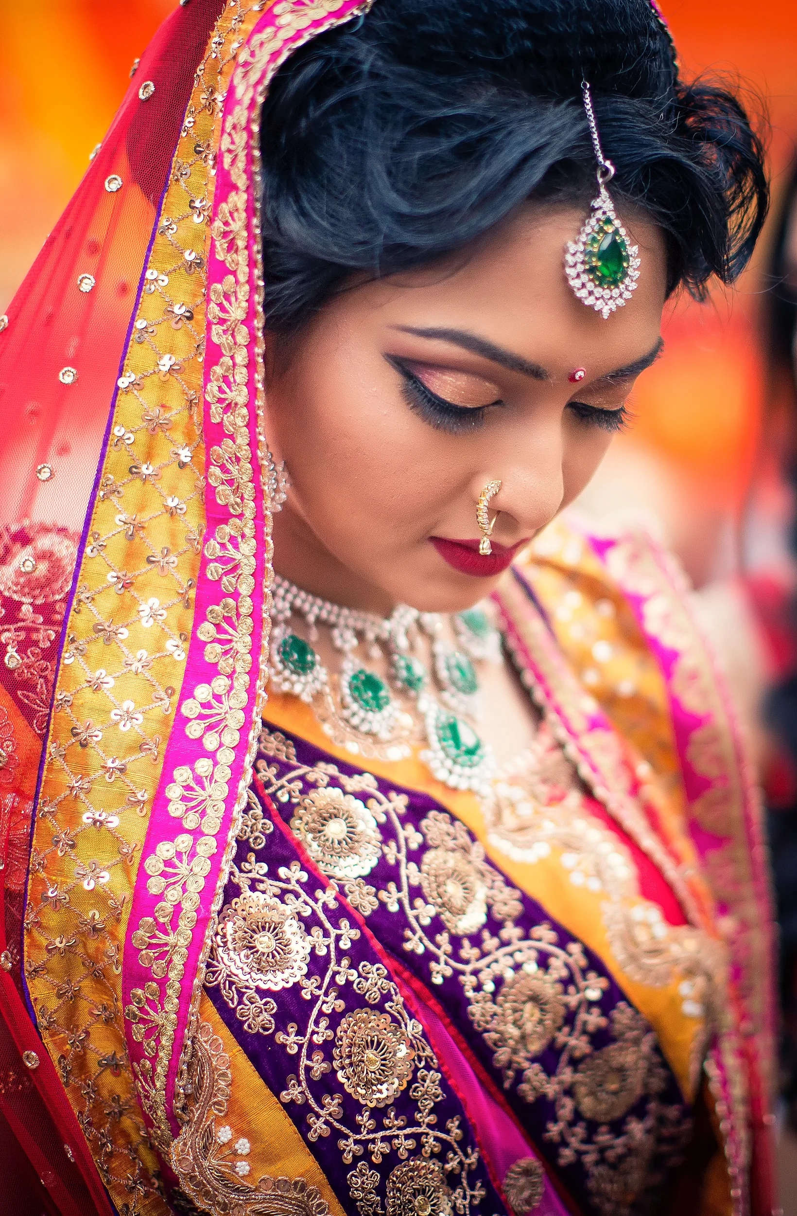 A woman dressed in traditional Indian attire with intricate gold embroidery, wearing jewelry including a nose ring, necklace, earrings, and a headpiece. She has her eyes closed and is looking downward, with a colorful orange, pink, and purple backgro