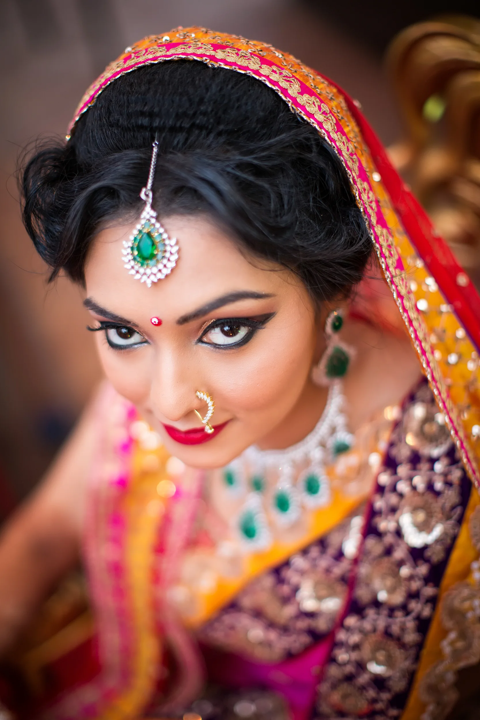Close-up of an Indian bride dressed in traditional attire, wearing jewelry and makeup, looking up at the camera with a smile.