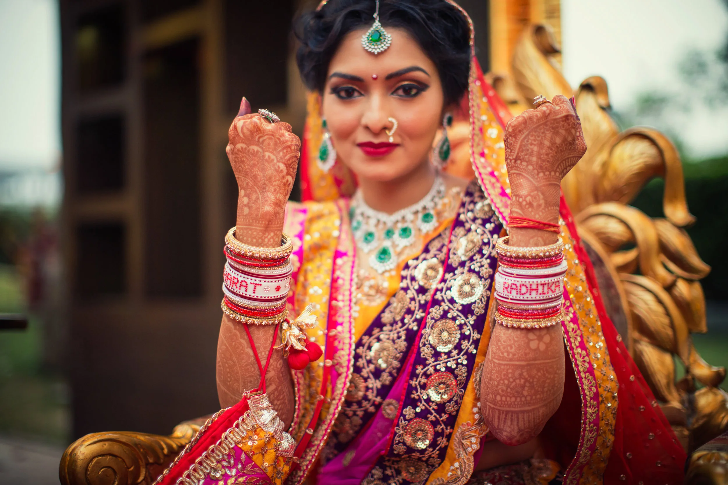 A woman dressed in traditional Indian bridal attire, sitting on a carved wooden chair, showing off her detailed henna-covered hands and jewelry, with a confident smile and fists raised.