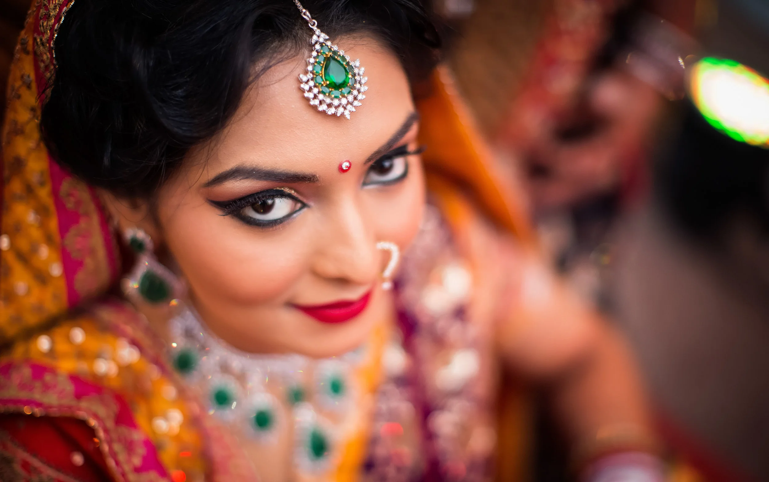 Close-up of an Indian bride with colorful traditional attire, jewelry, and makeup, looking up at the camera.
