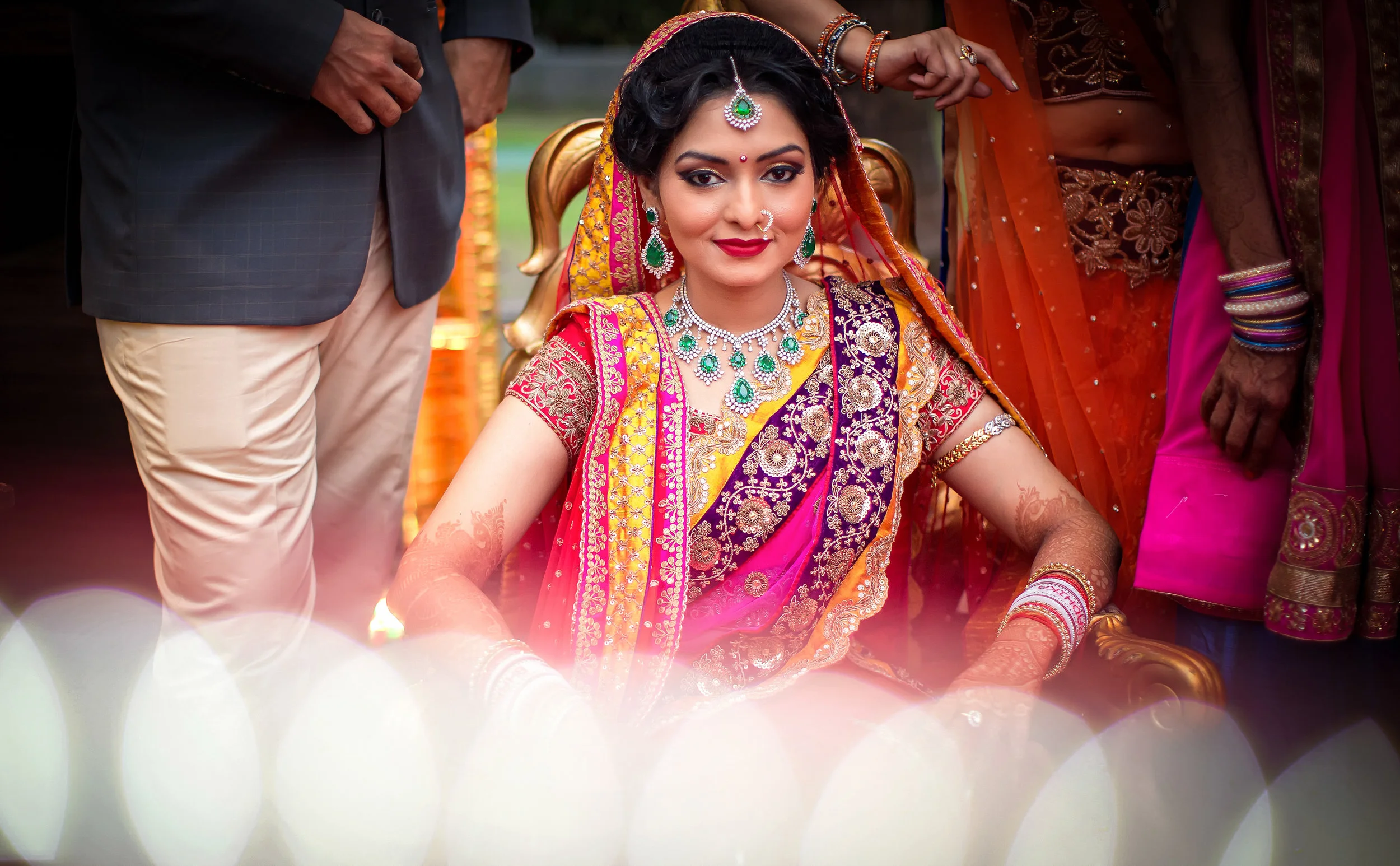 Indo-Western bride wearing traditional jewelry and colorful saree, sitting on a decorated chair during wedding ceremony, surrounded by family members.