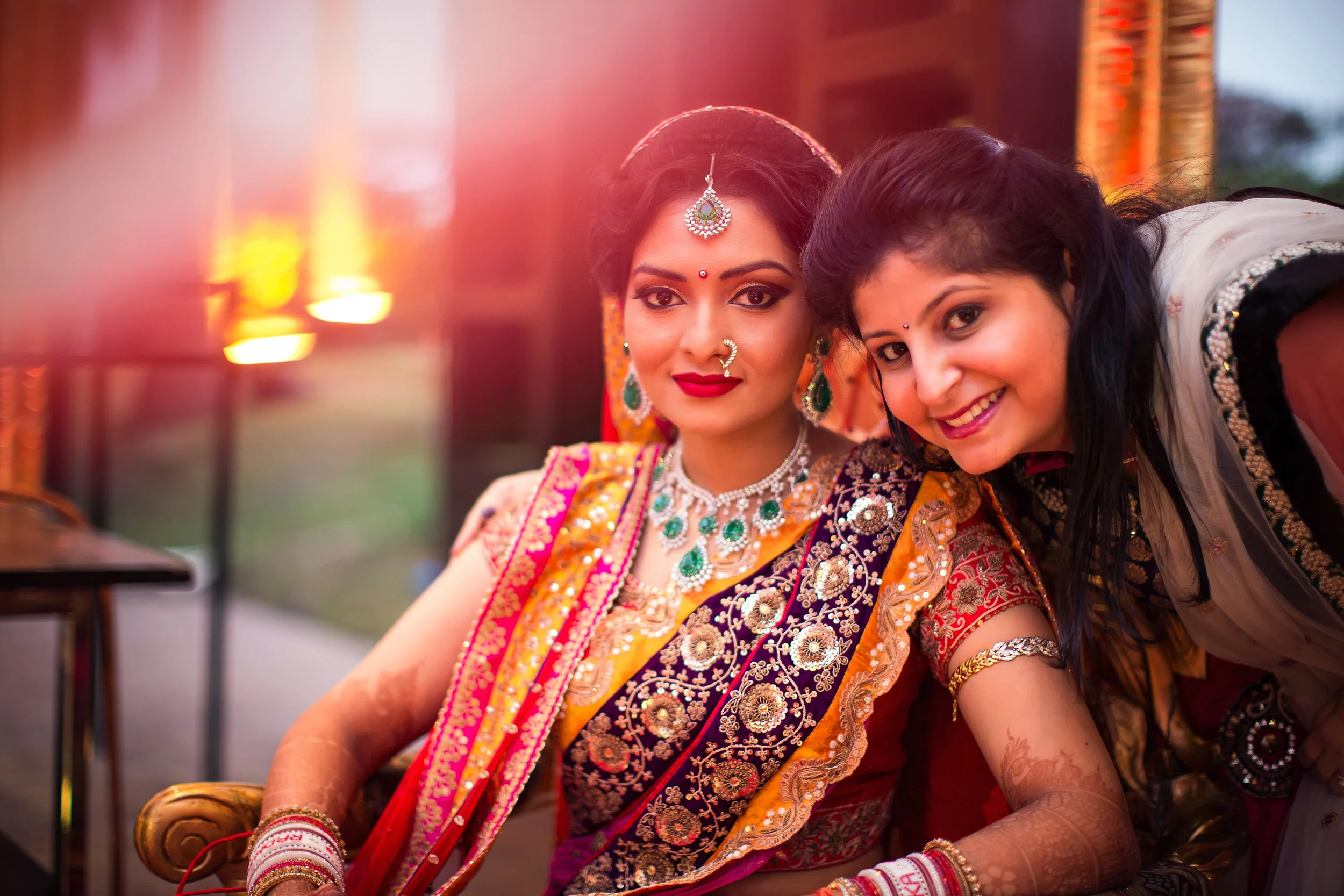 A woman wearing traditional Indian bridal attire with jewelry, sitting outdoors during sunset, with another woman leaning in beside her and smiling.