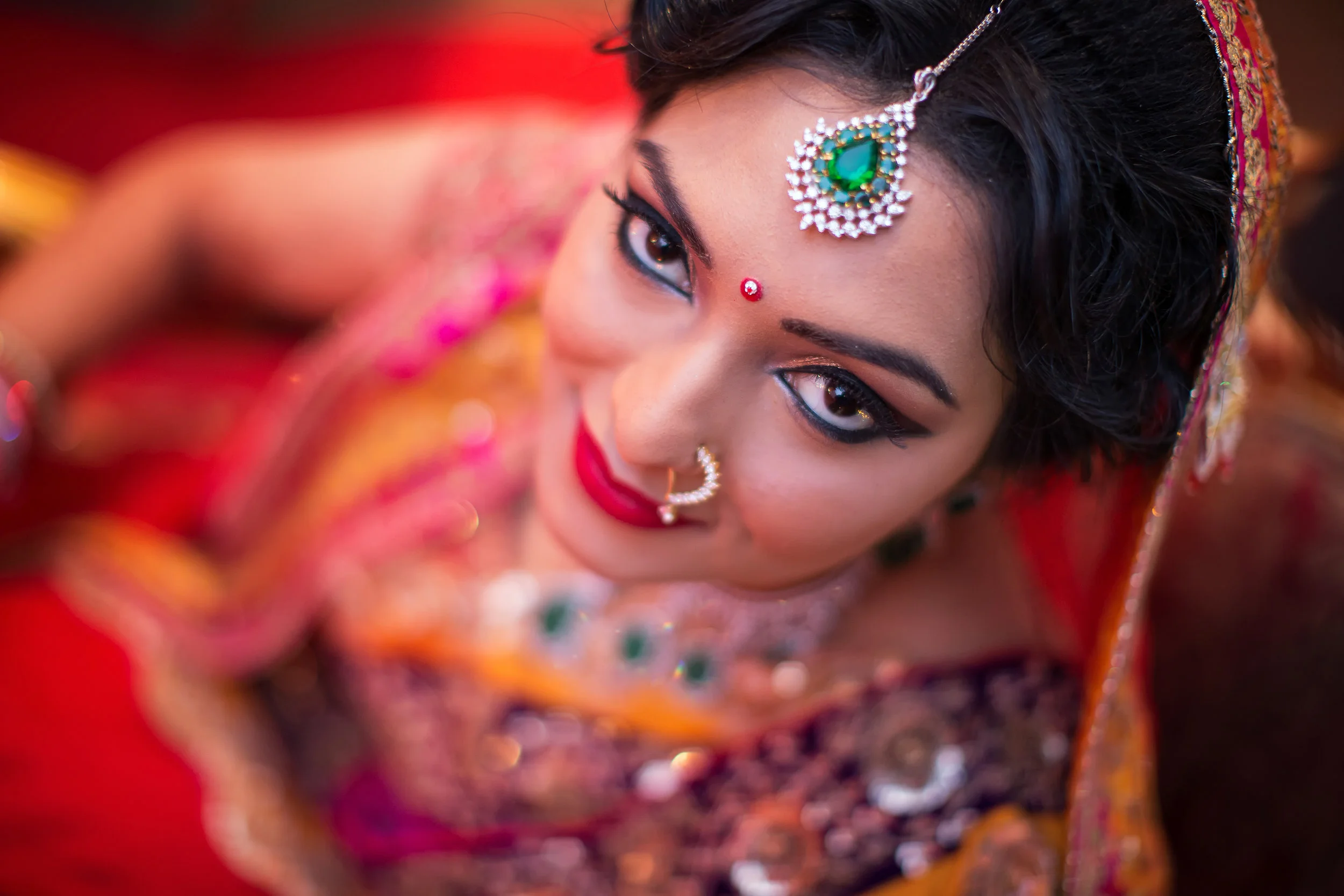 Close-up of Indian bride in traditional colorful attire with jewelry, eye makeup, and red bindi, looking upwards.