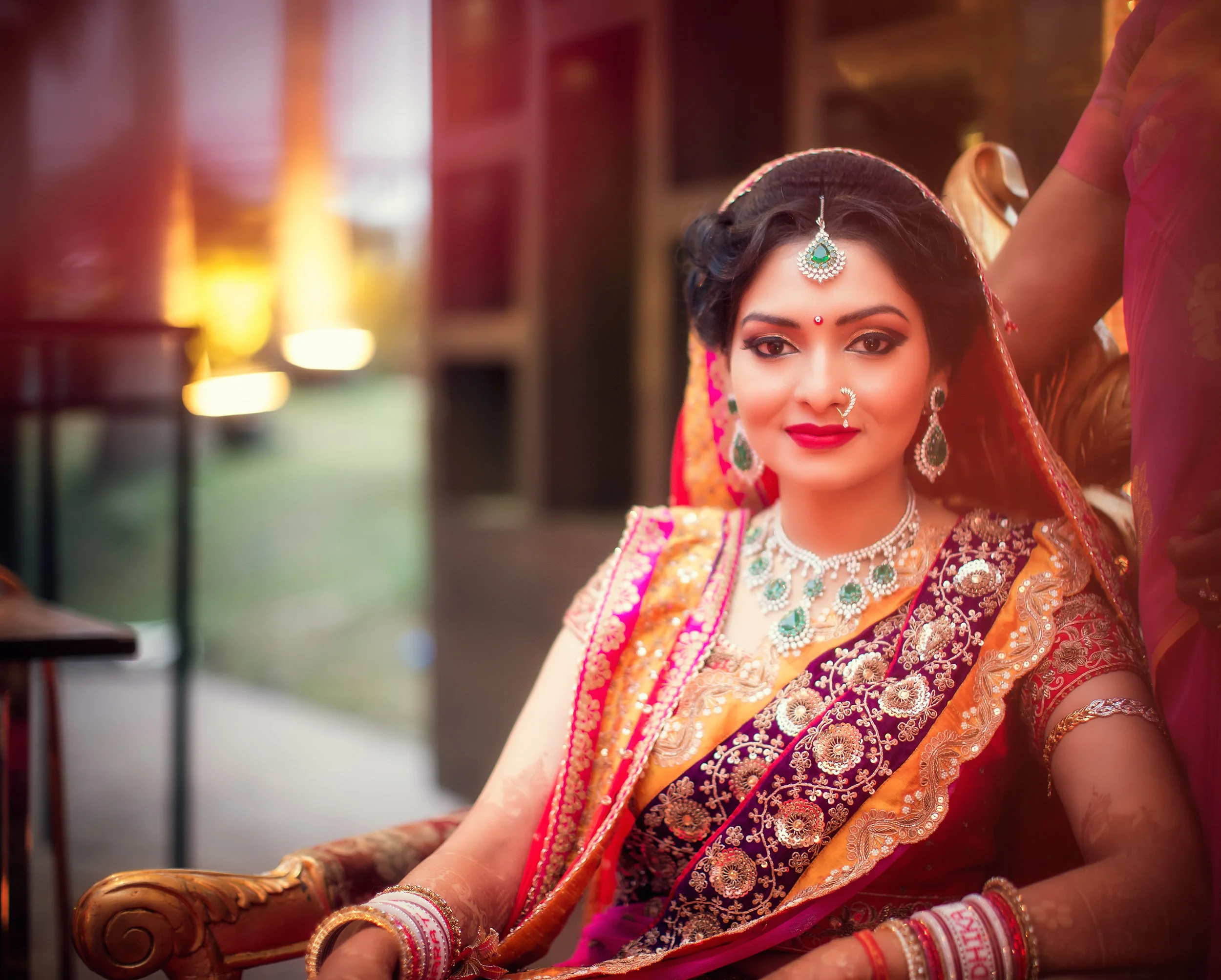 A woman dressed in traditional Indian attire, wearing jewelry and makeup, sitting on a wooden chair.