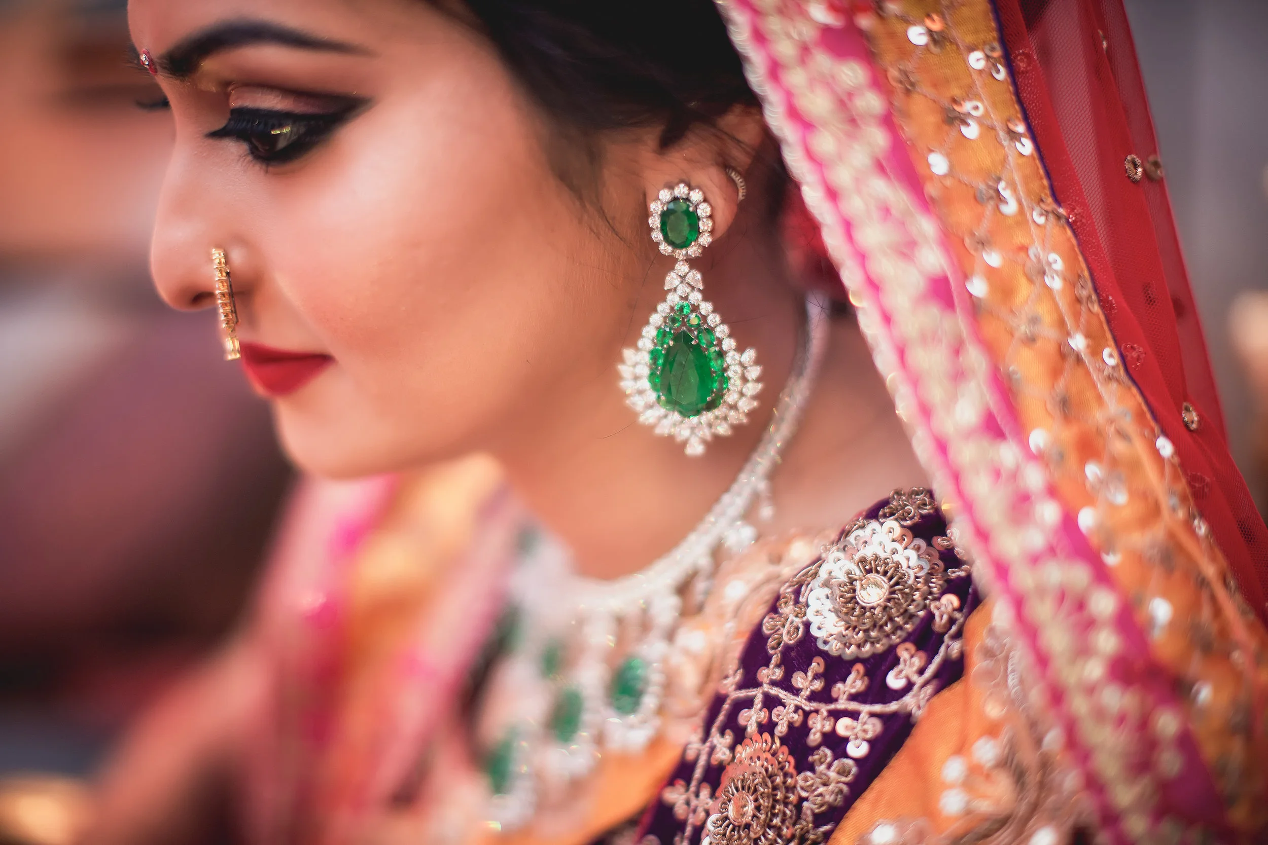 Close-up of a woman wearing traditional Indian jewelry and colorful embroidered attire, with intricate earrings, nose ring, and vibrant fabric with sequins.