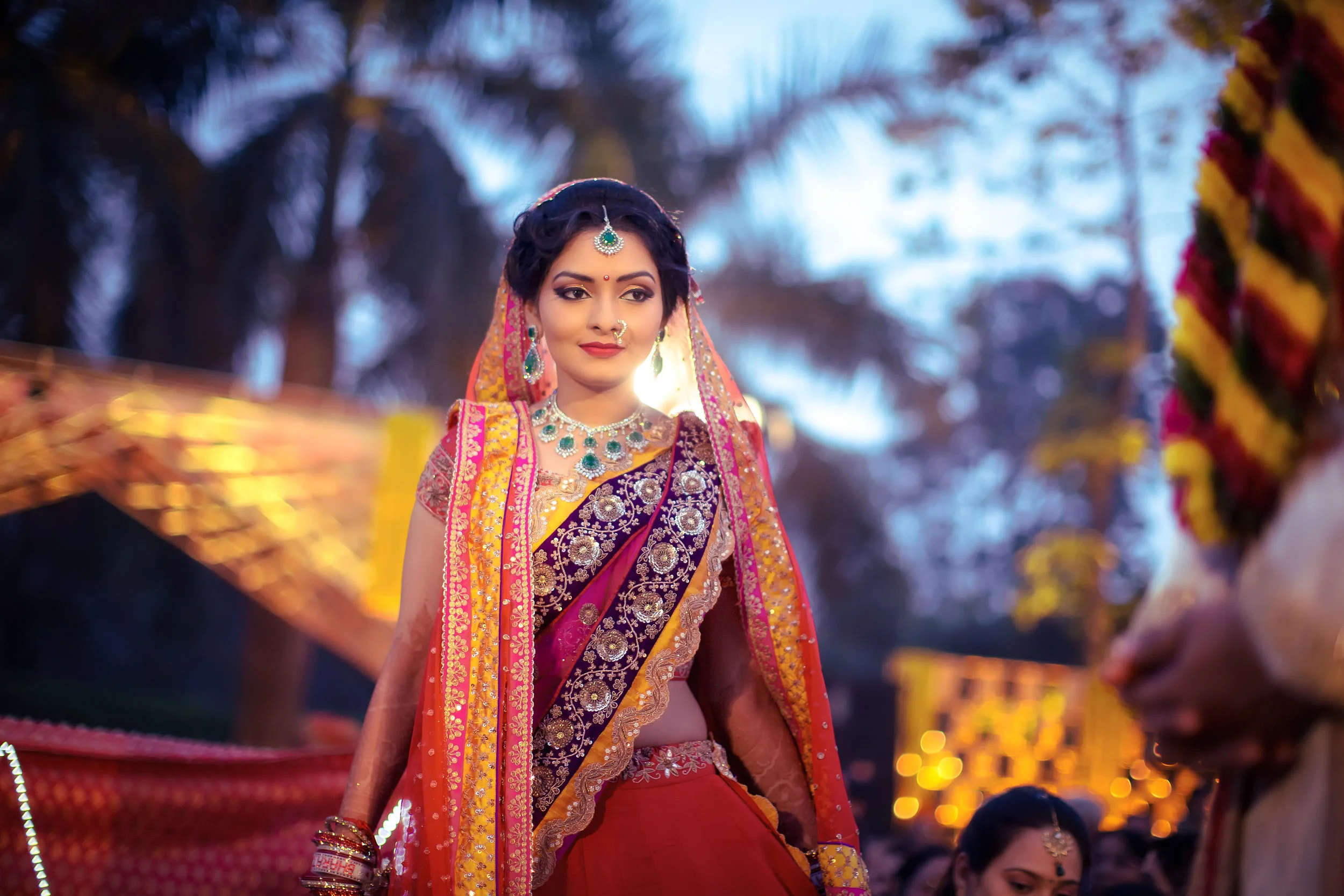 A woman dressed in traditional Indian wedding attire, wearing a richly embroidered saree in shades of purple, with jewelry including a necklace, earrings, and a nose ring, stands outdoors during sunset with trees in the background.
