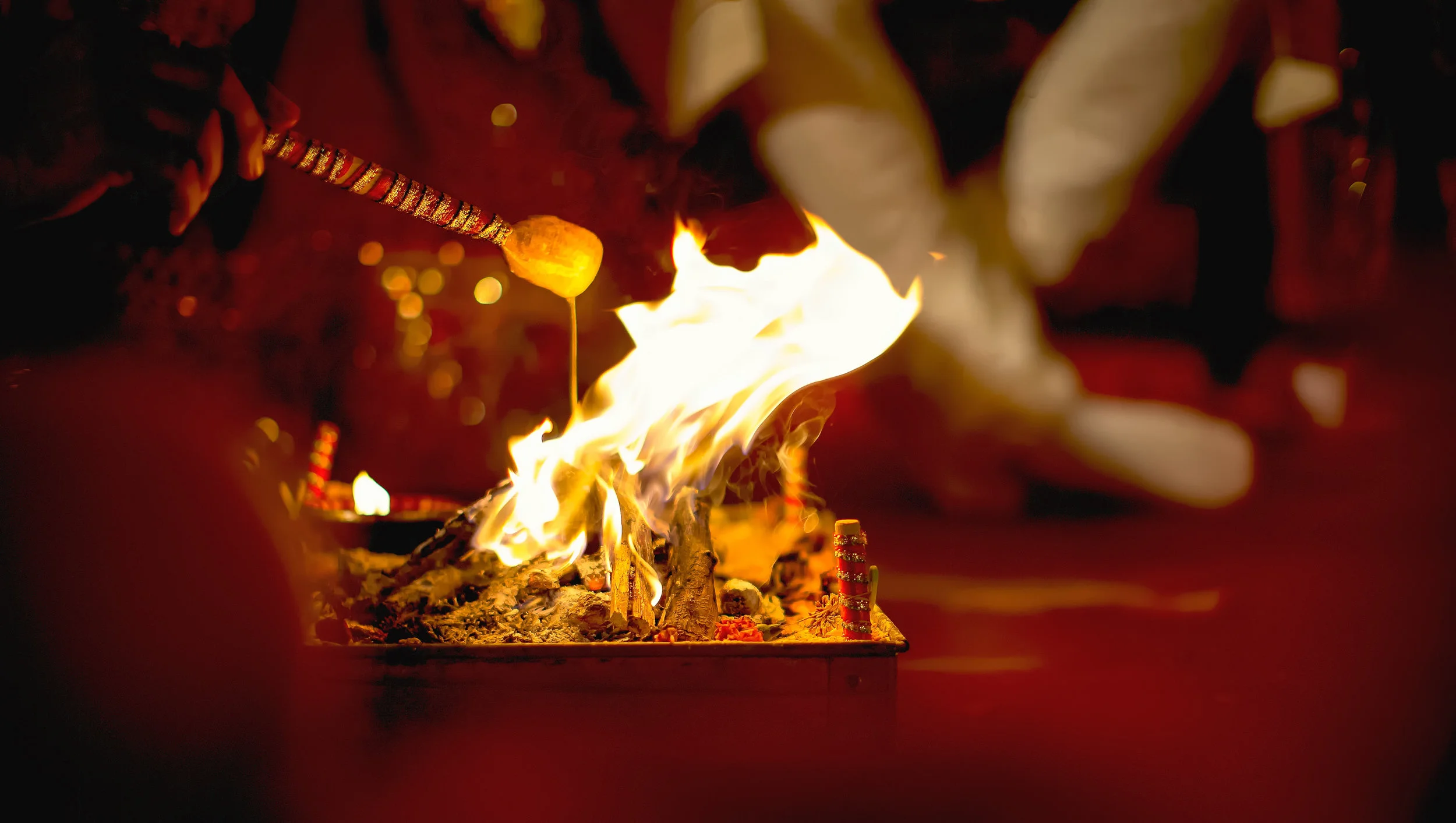 A person blows a flame on a small ceremonial incense fire with flaming sticks, in a dark setting with a warm glow.