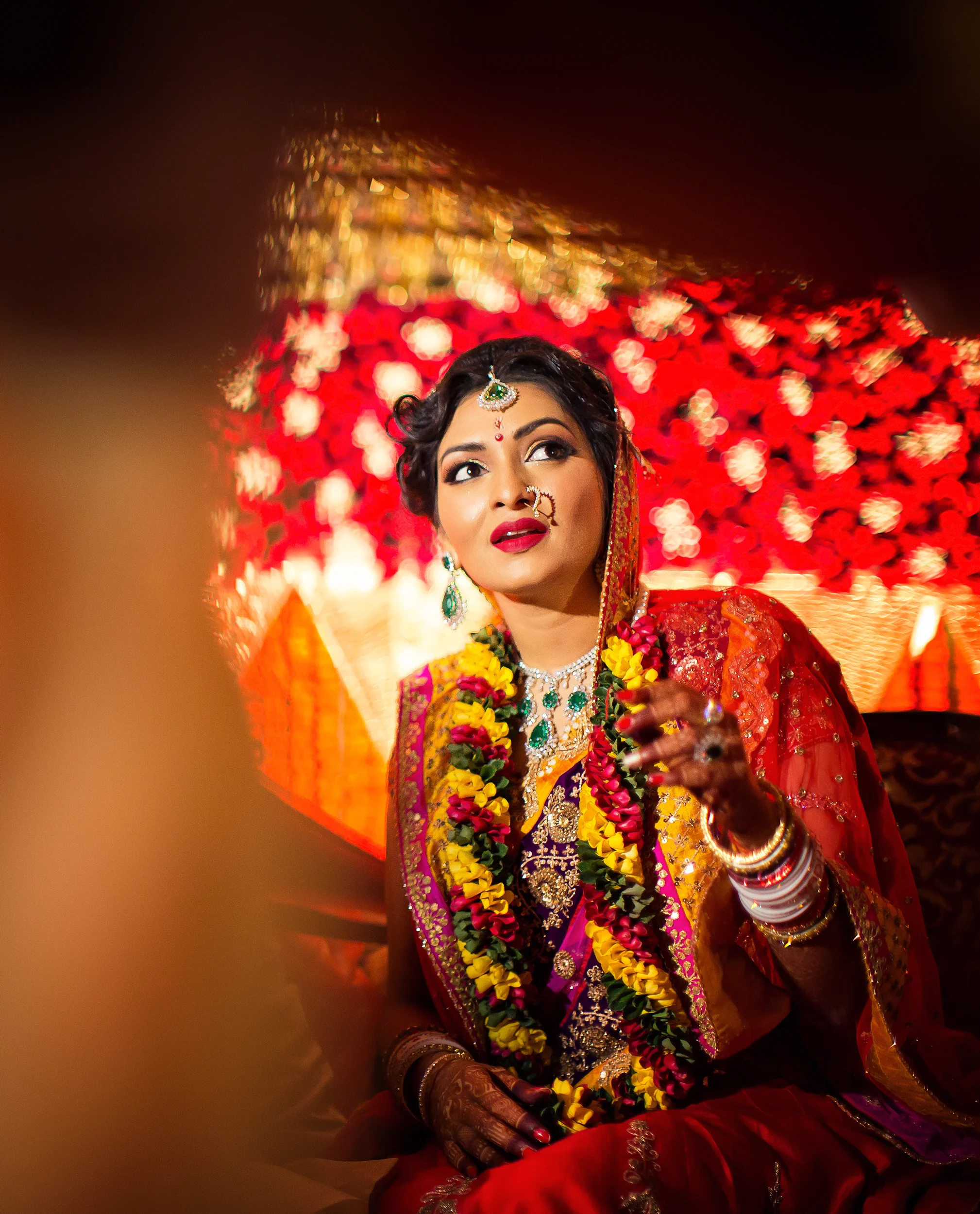 A woman dressed in traditional Indian wedding attire, wearing jewelry and a colorful garland, seated against a backdrop of red roses and golden decorations.