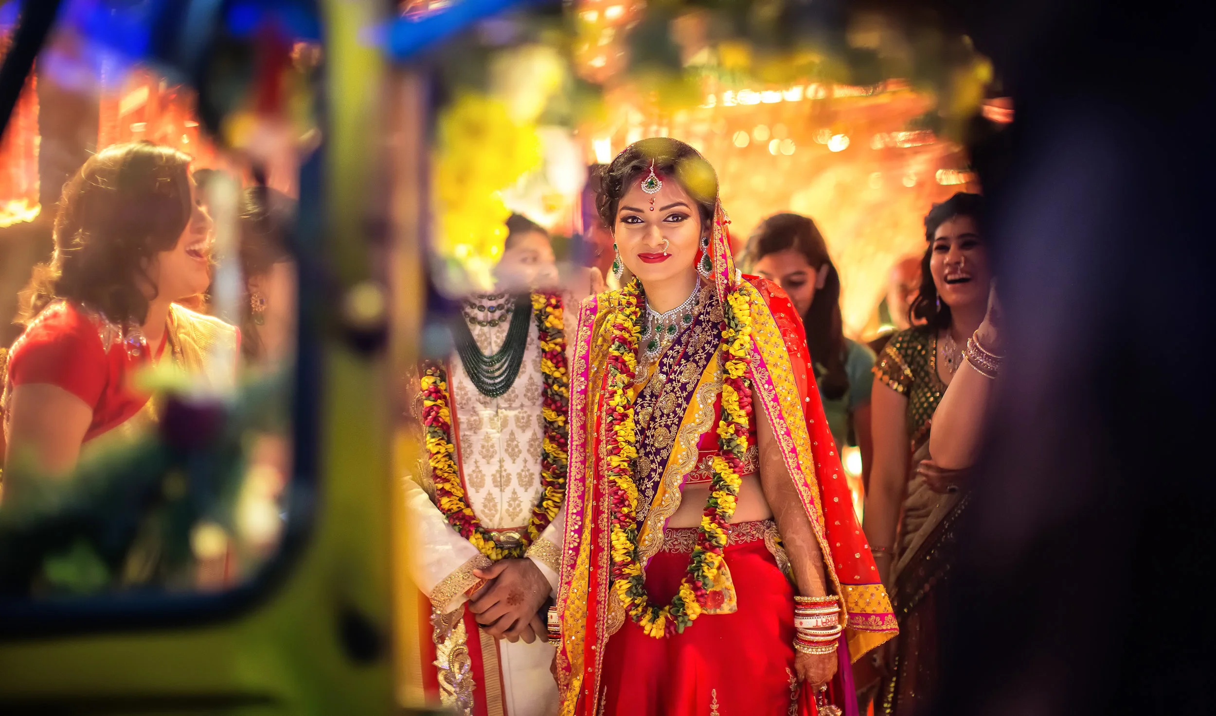 Indian wedding celebration with women in colorful traditional attire and floral garlands, festive lights in the background.