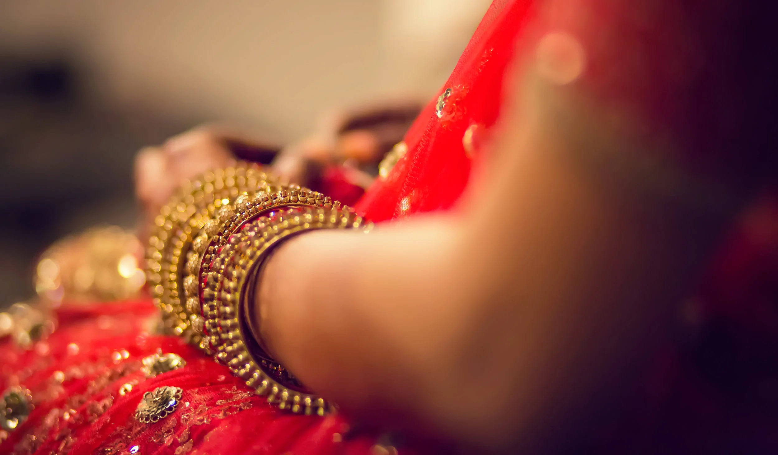 Close-up of gold bangles worn on a woman's wrist, with a red fabric and intricate embroidery.
