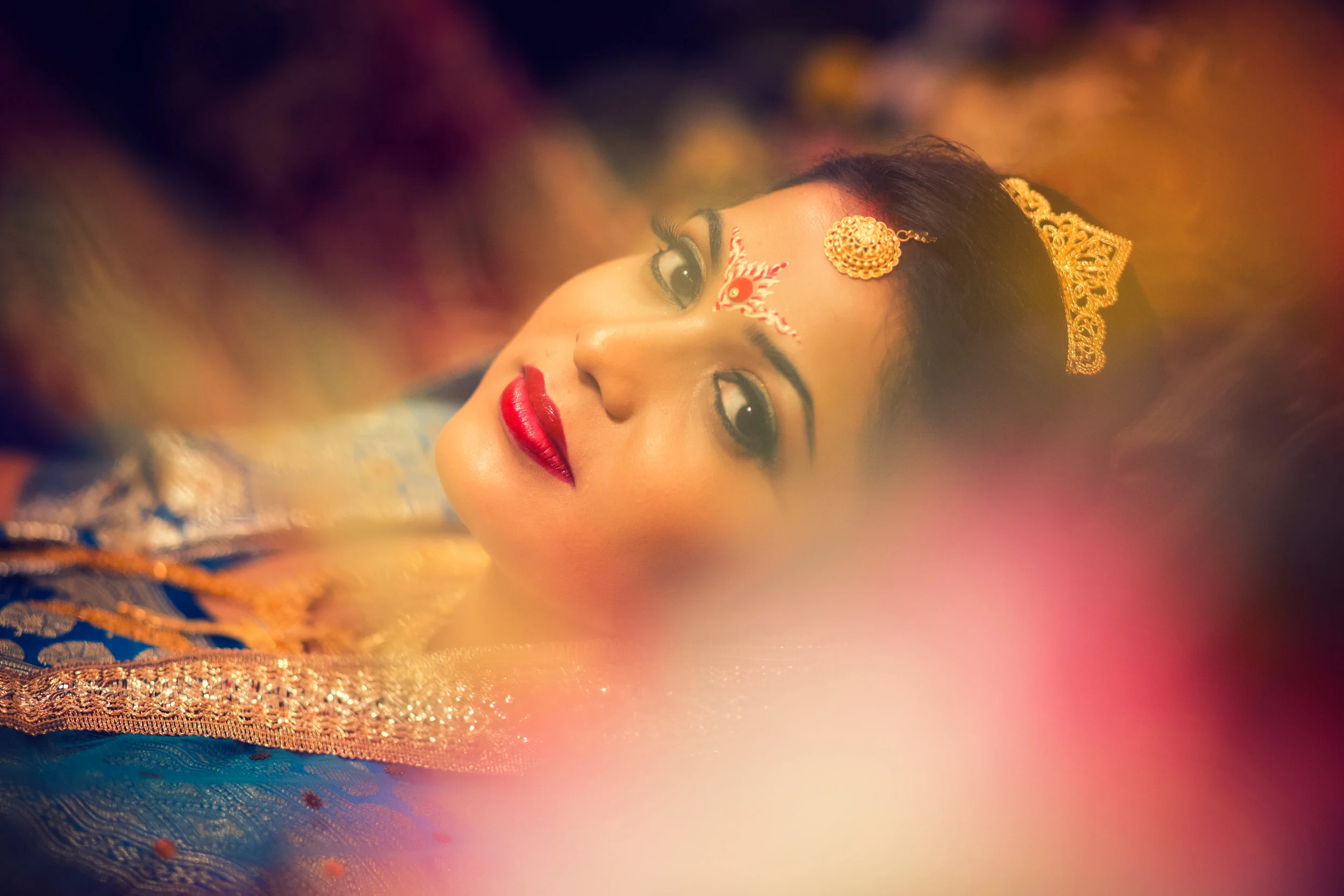Close-up of a woman with traditional Indian jewelry and makeup, lying down, with colorful and blurred foreground.