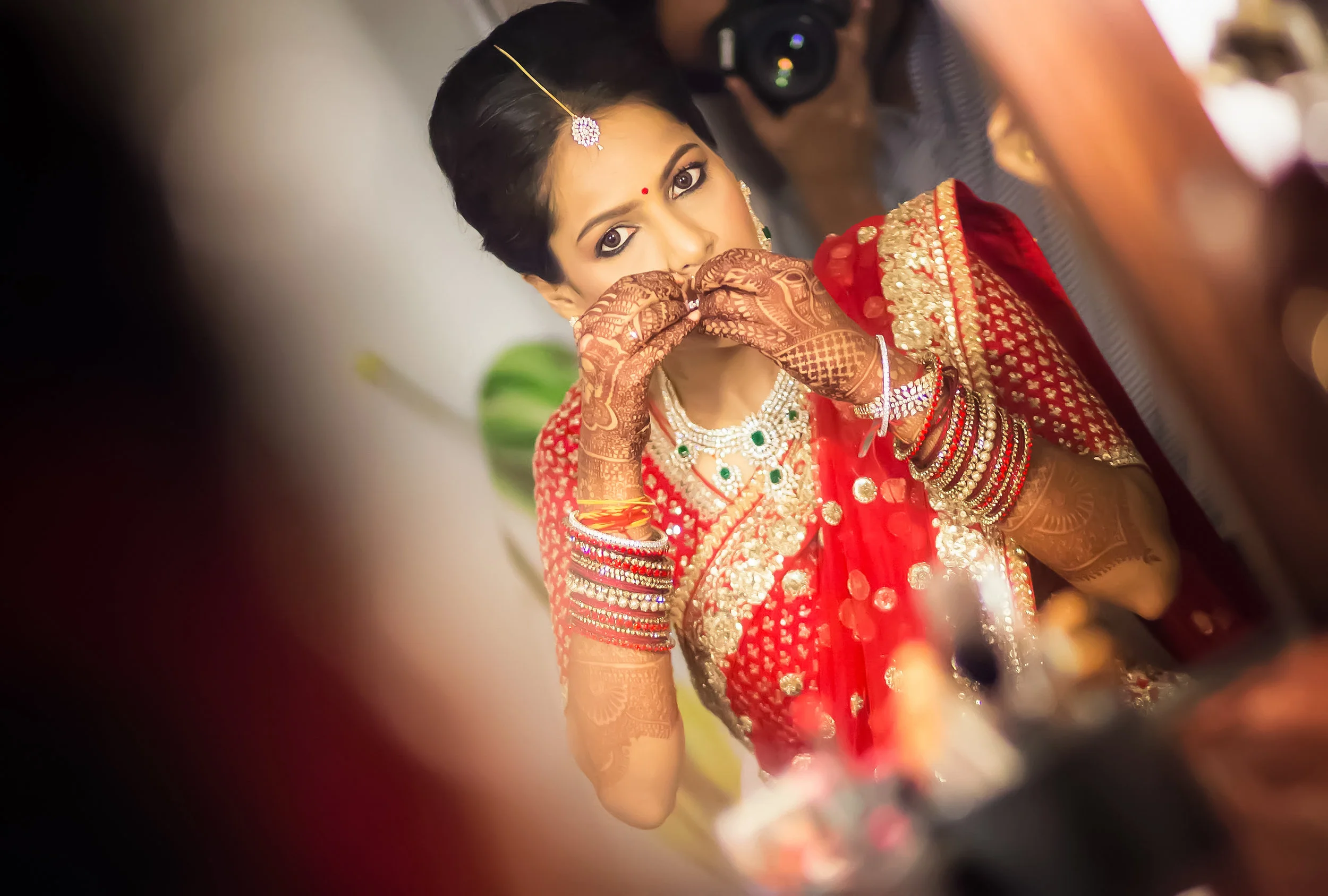 A woman dressed in traditional Indian bridal attire, wearing a red saree with gold embroidery, jewelry, and henna tattoos, is covering her mouth with her hands during her wedding ceremony.