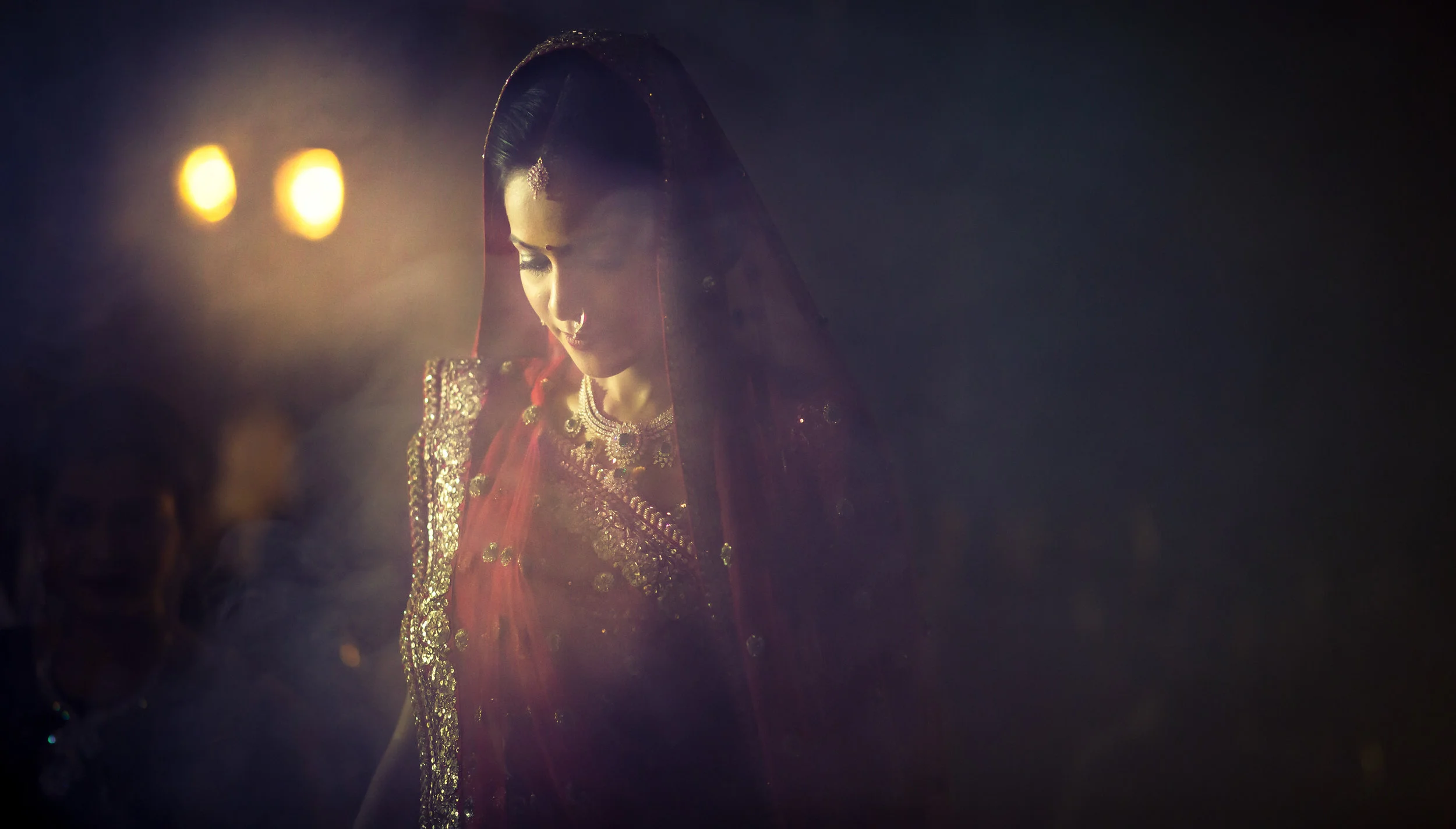 A woman dressed in traditional Indian attire with jewelry, wearing a red saree or lehenga, and a veil, standing in a dimly lit setting with soft lighting.