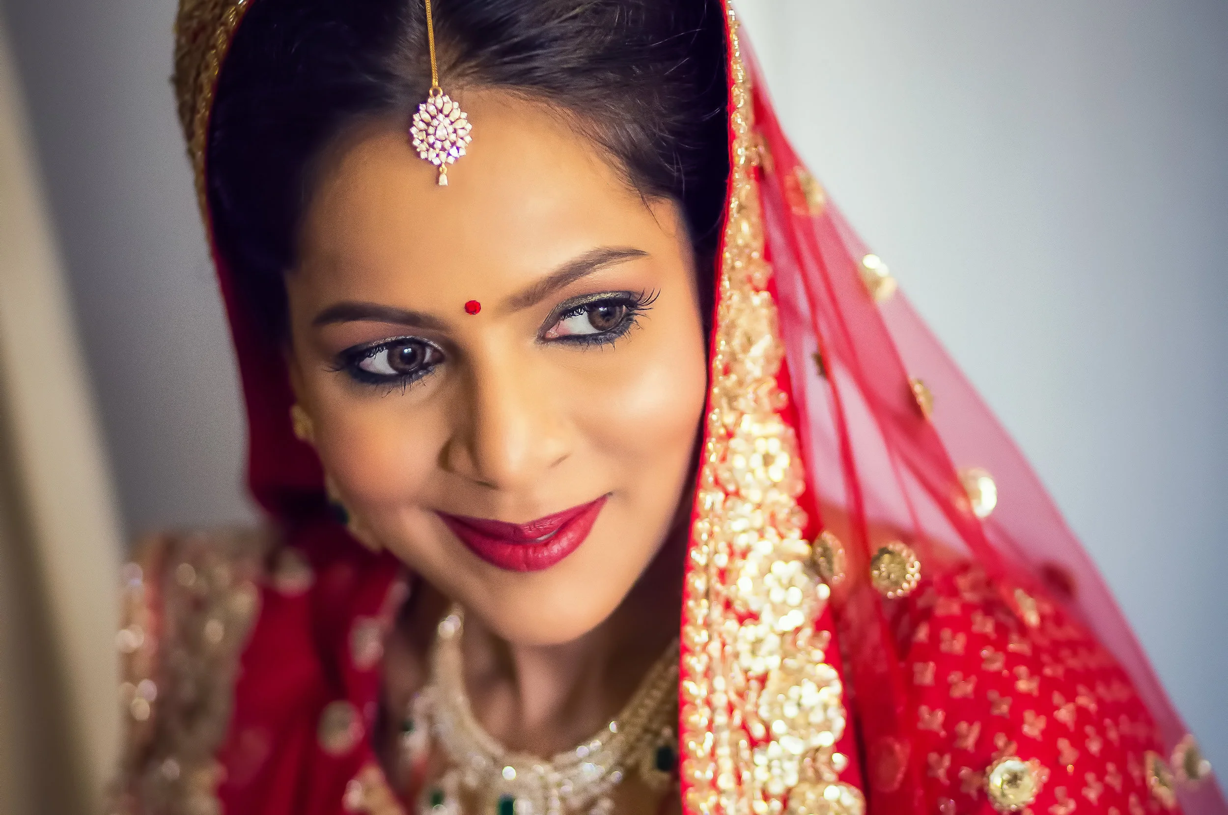 Close-up of a woman dressed in traditional Indian wedding attire with ornate jewelry and a red veil, smiling.
