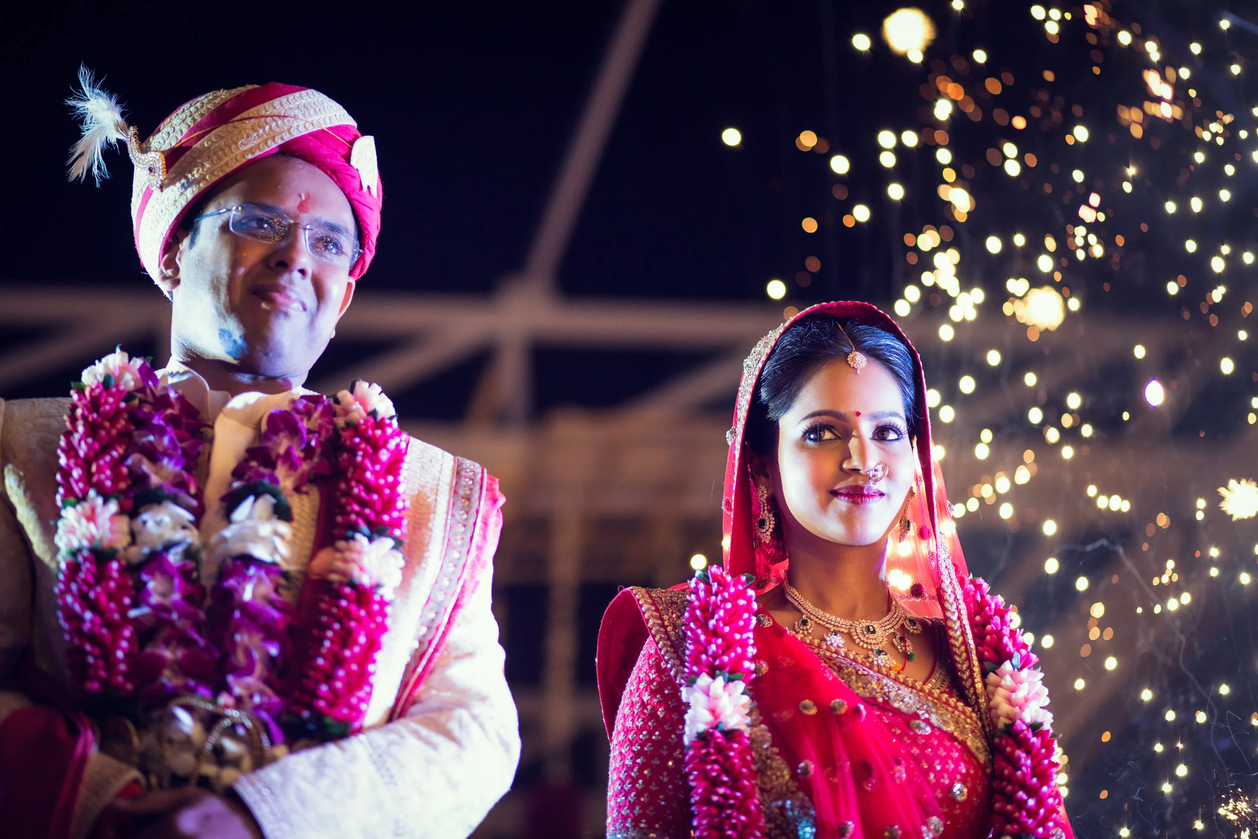 Indian bride and groom in traditional wedding attire during nighttime celebration, with fireworks in the background.