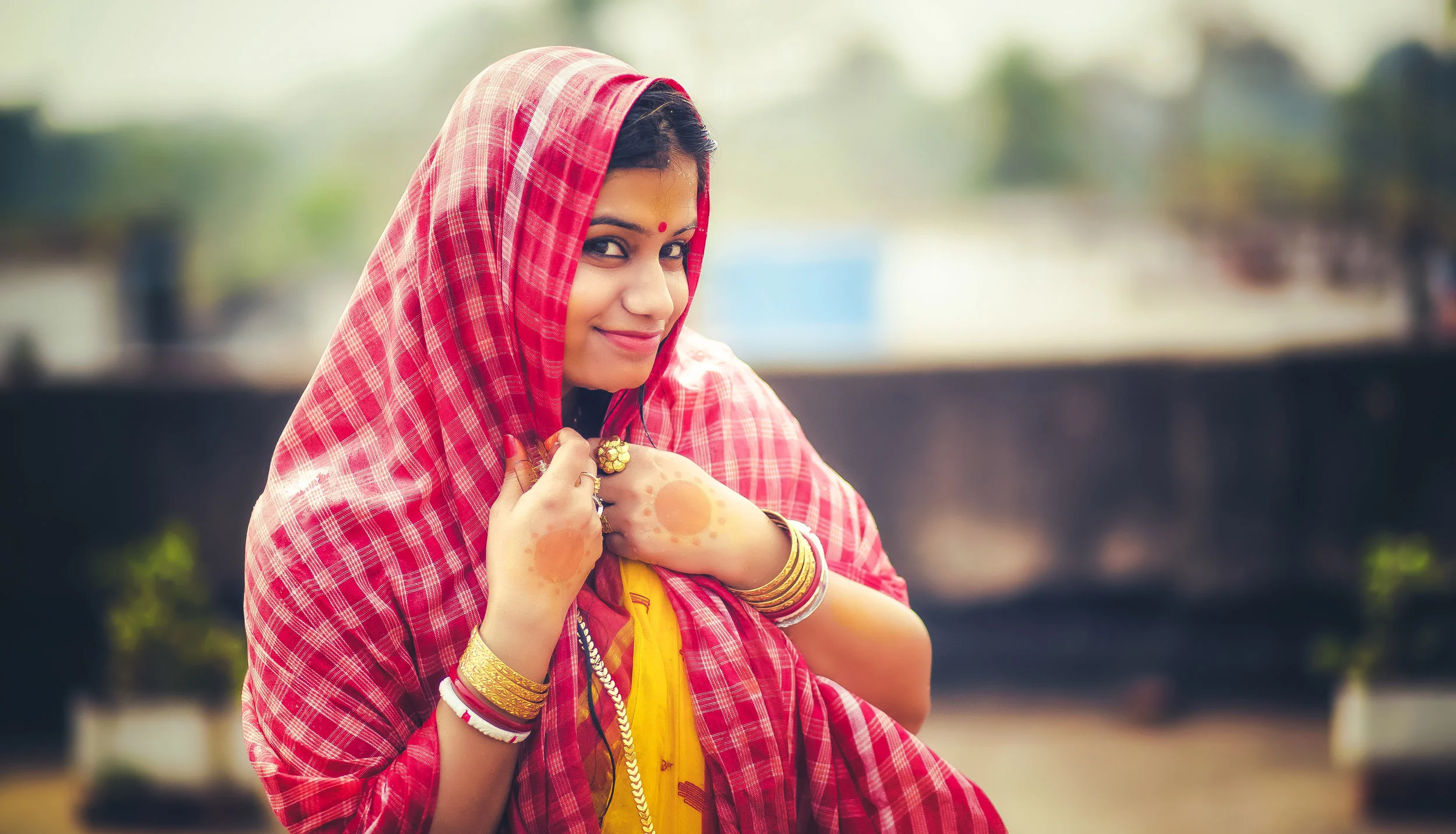 A woman dressed in traditional Indian attire, wearing a red and yellow sari, adorned with bangles, rings, and henna on her hands, is smiling and holding her sari with one hand against an outdoor background.